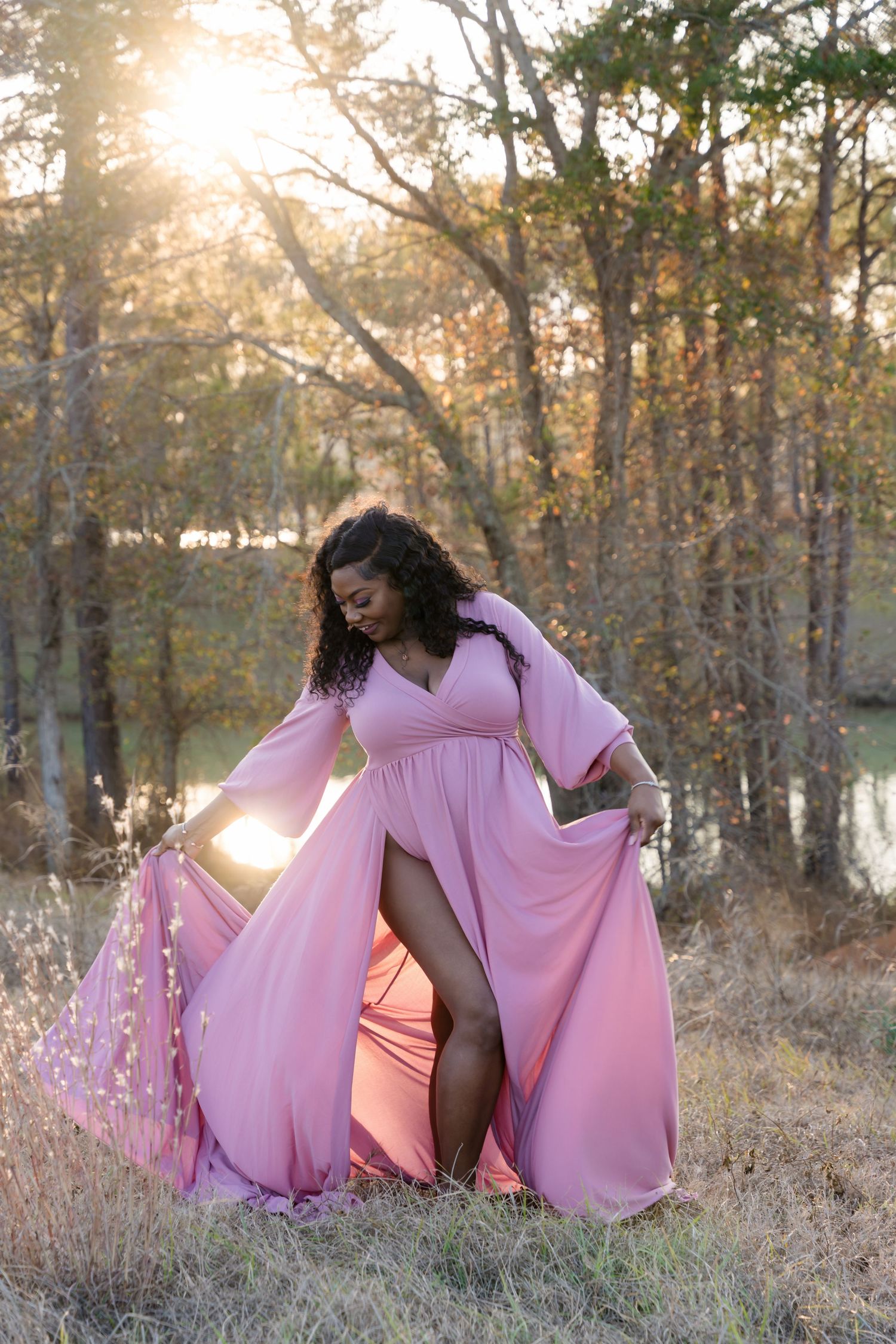 Woman in flowing pink gown poses gracefully outdoors at golden hour among autumn trees near a pond.