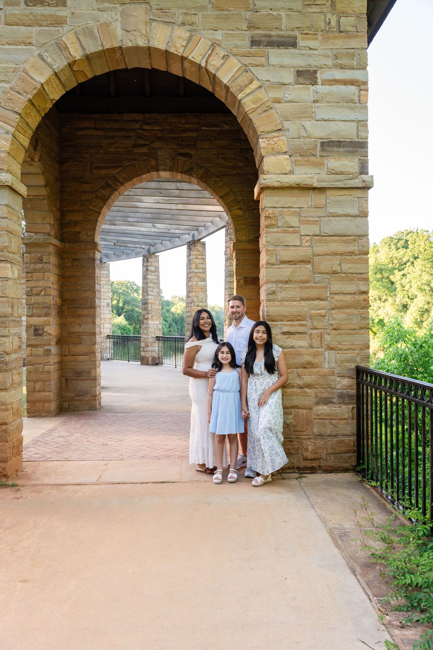 Family of four in light summer outfits posing together beneath a stone archway with a scenic walkway and trees behind them.
