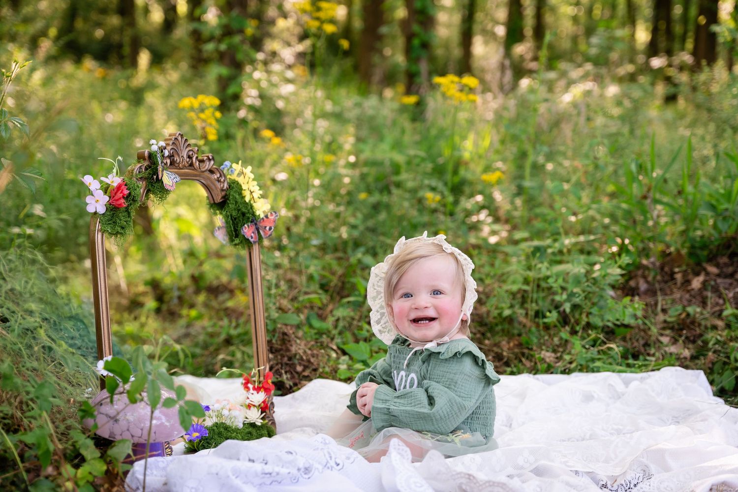 Smiling baby girl in green dress and lace bonnet sitting on white blanket in lush forest garden setting.