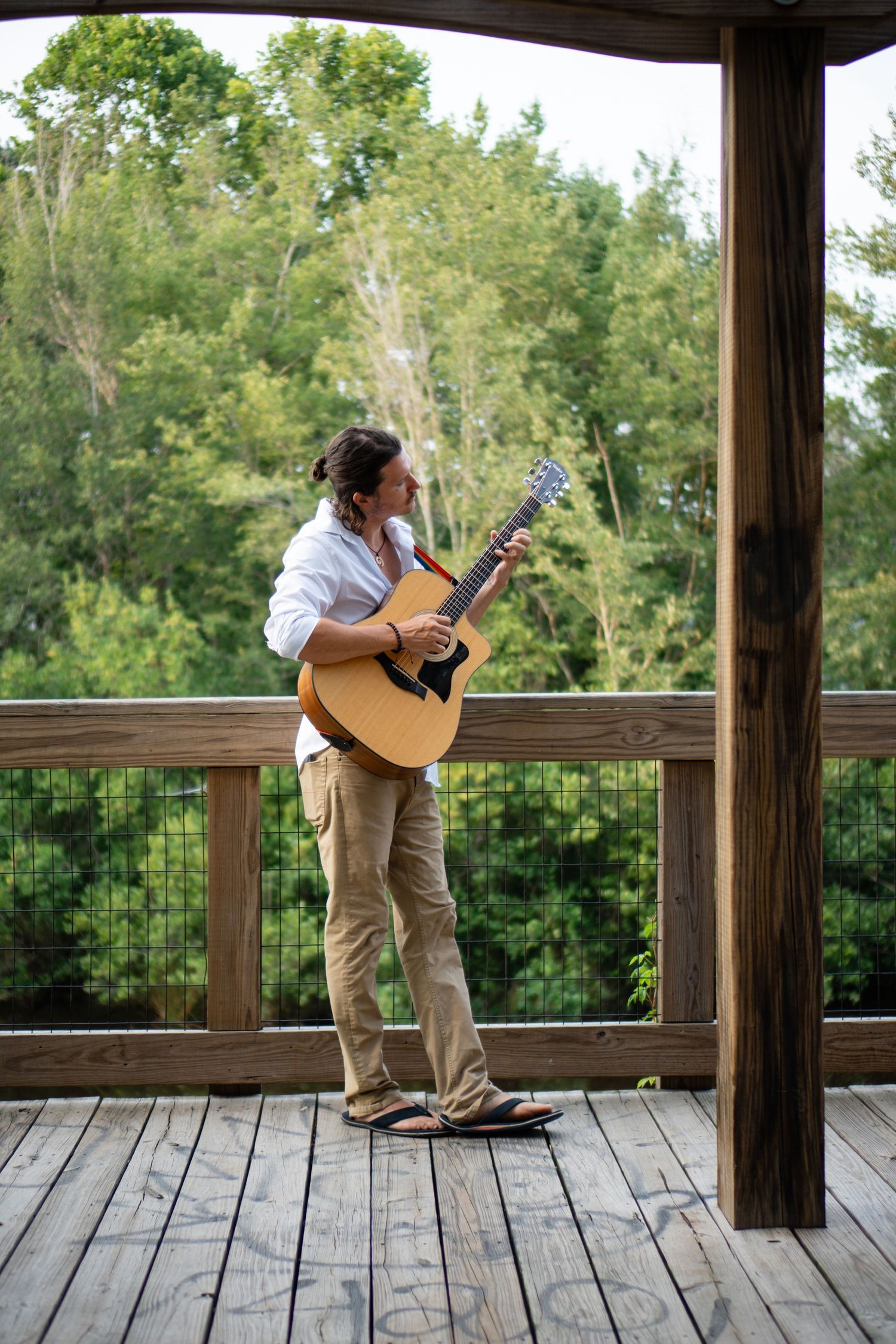 Musician performing with acoustic guitar on wooden deck overlooking forest landscape.