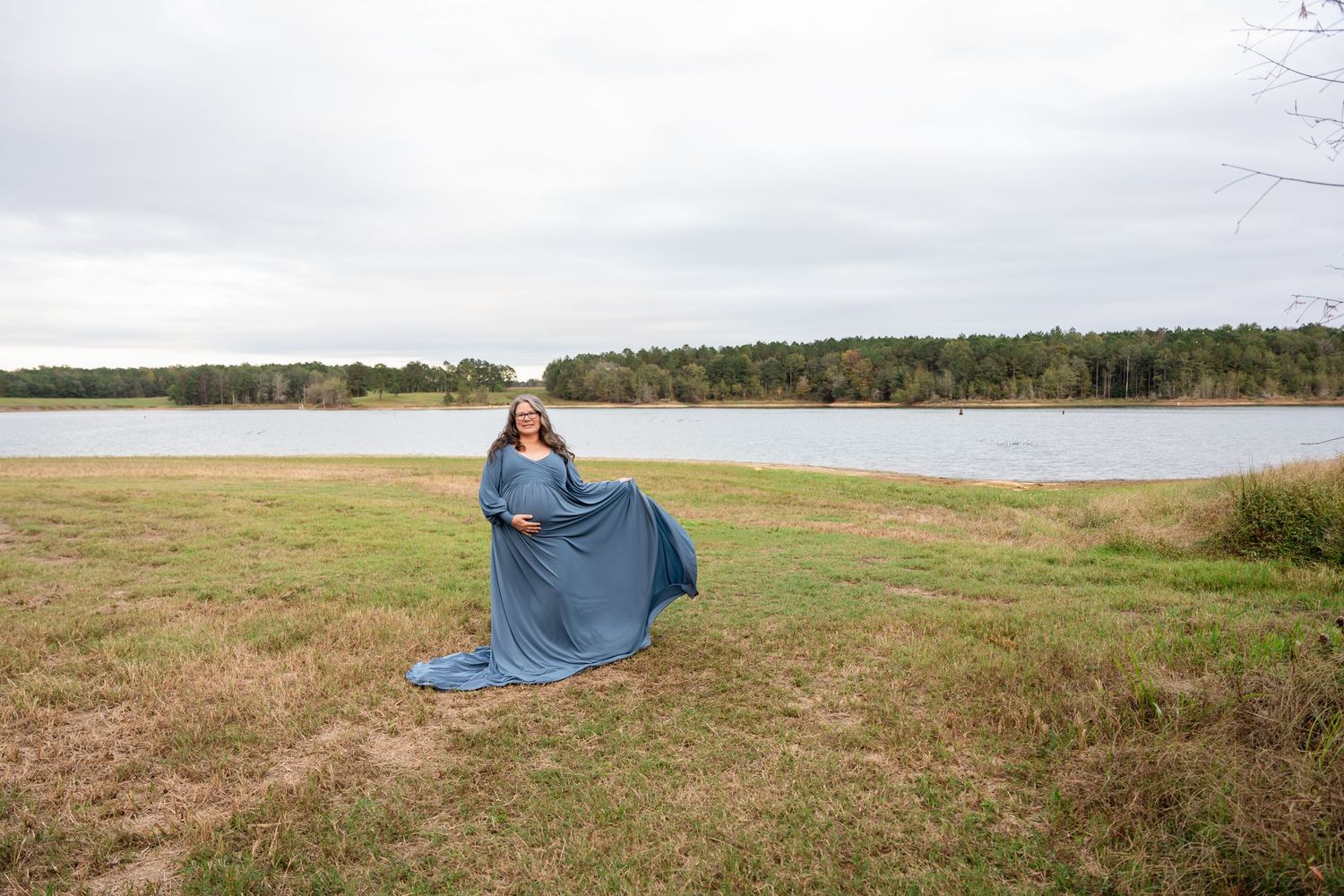 Woman in blue dress twirls gracefully against lakeside landscape on overcast day.