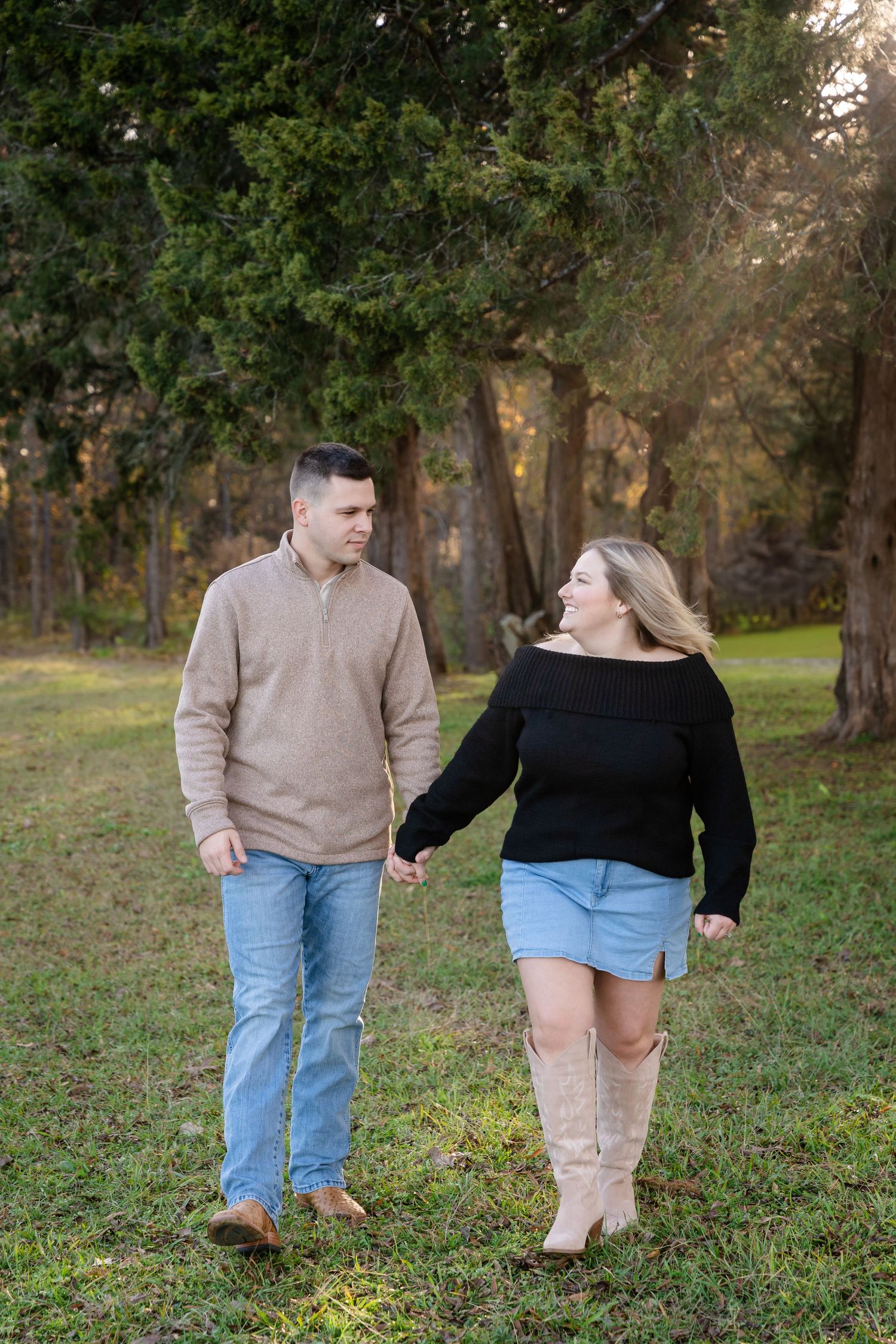 Couple walking hand in hand through grass wearing casual fall attire.