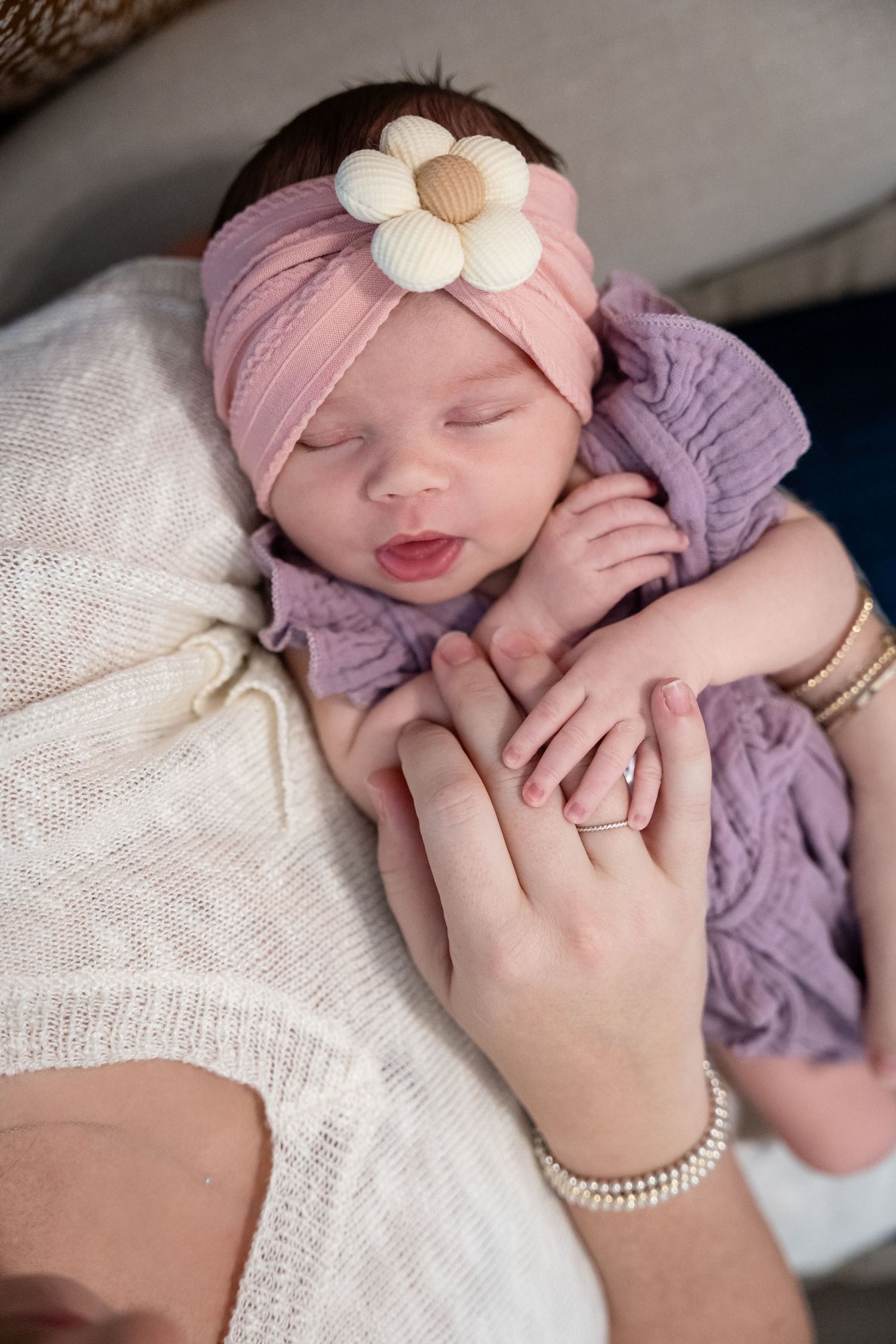 Sleeping newborn baby girl in purple knit wrap and pink floral headband cradled in mother's hands.
