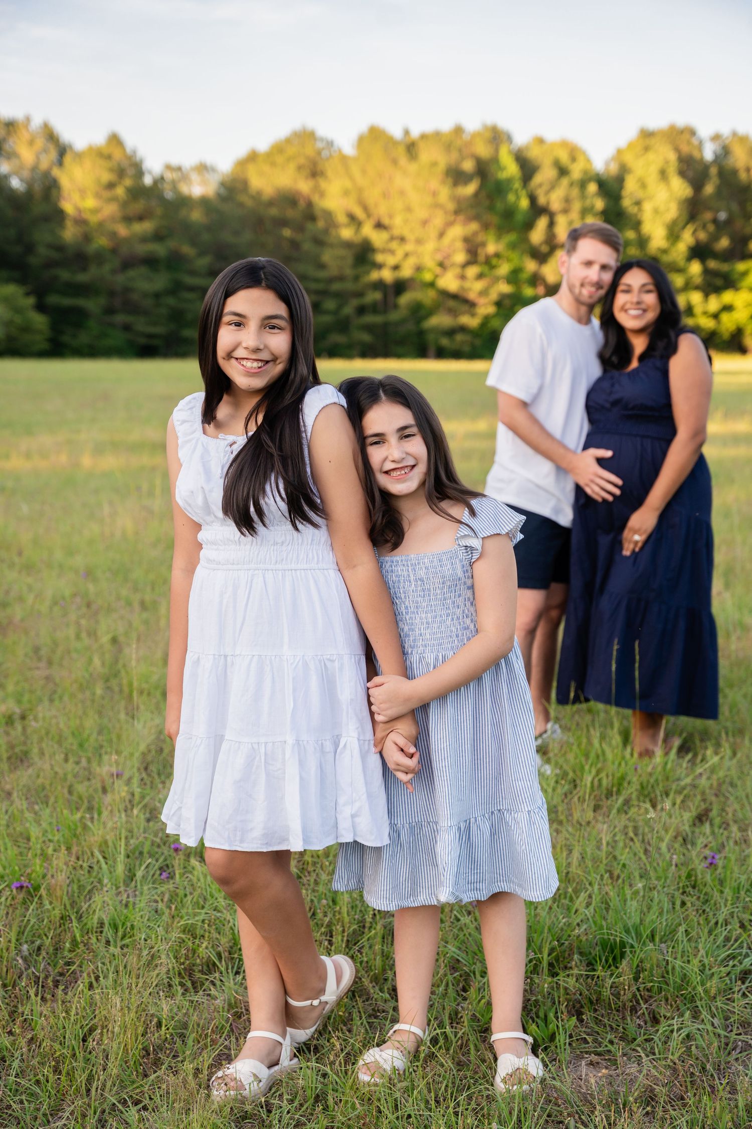 Two girls in white and blue dresses pose in a field with their parents standing behind them at sunset.