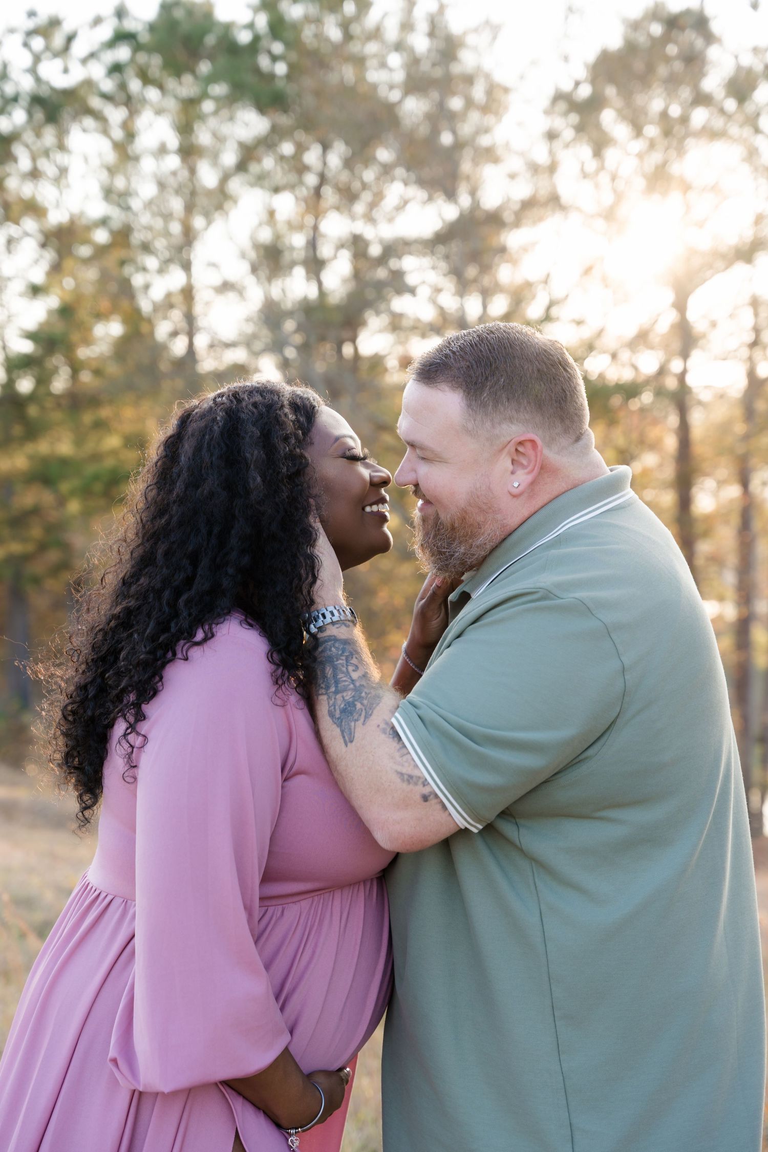 A couple shares a romantic moment outdoors at golden hour, woman in pink dress and man in green polo smiling at each other.