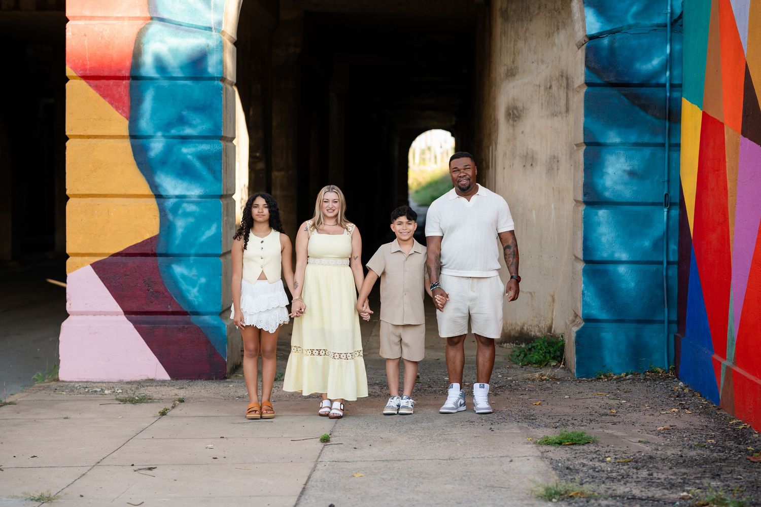 Family of four posing in front of colorful murals under a bridge on a sunny day.