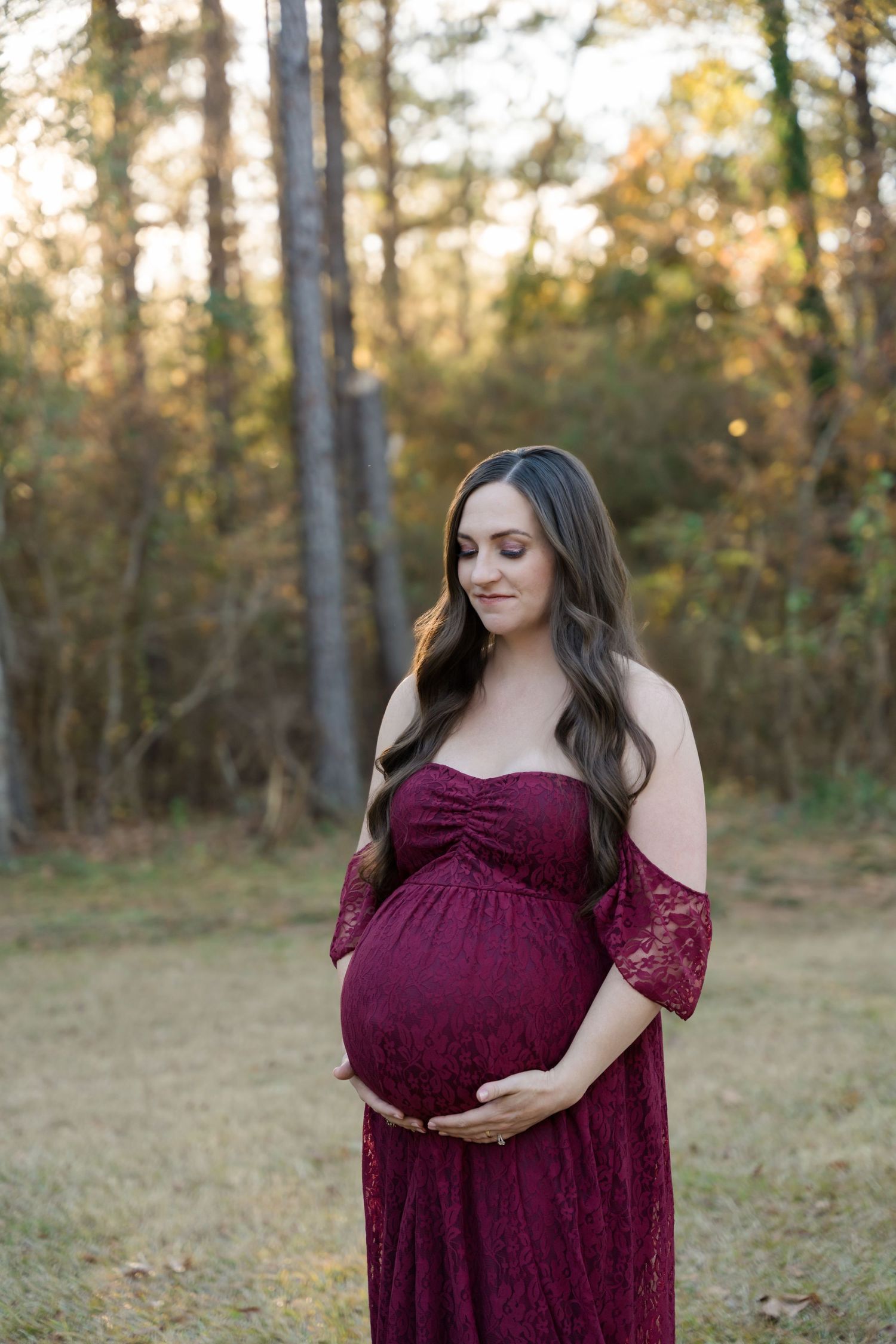 A pregnant woman in a burgundy lace gown stands in a sunlit forest clearing during golden hour.