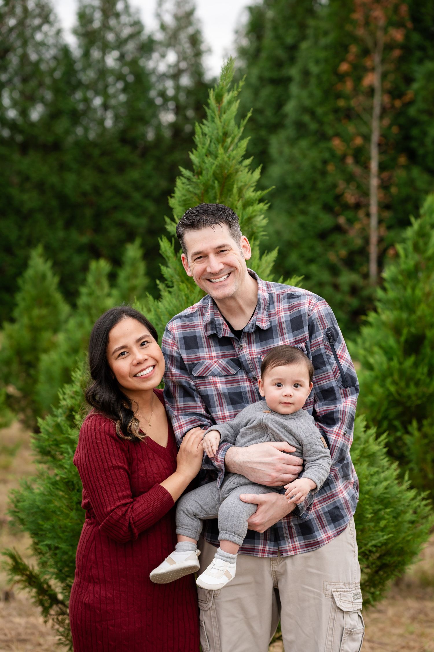 Happy family poses for portraits in front of evergreen trees during outdoor photo session.