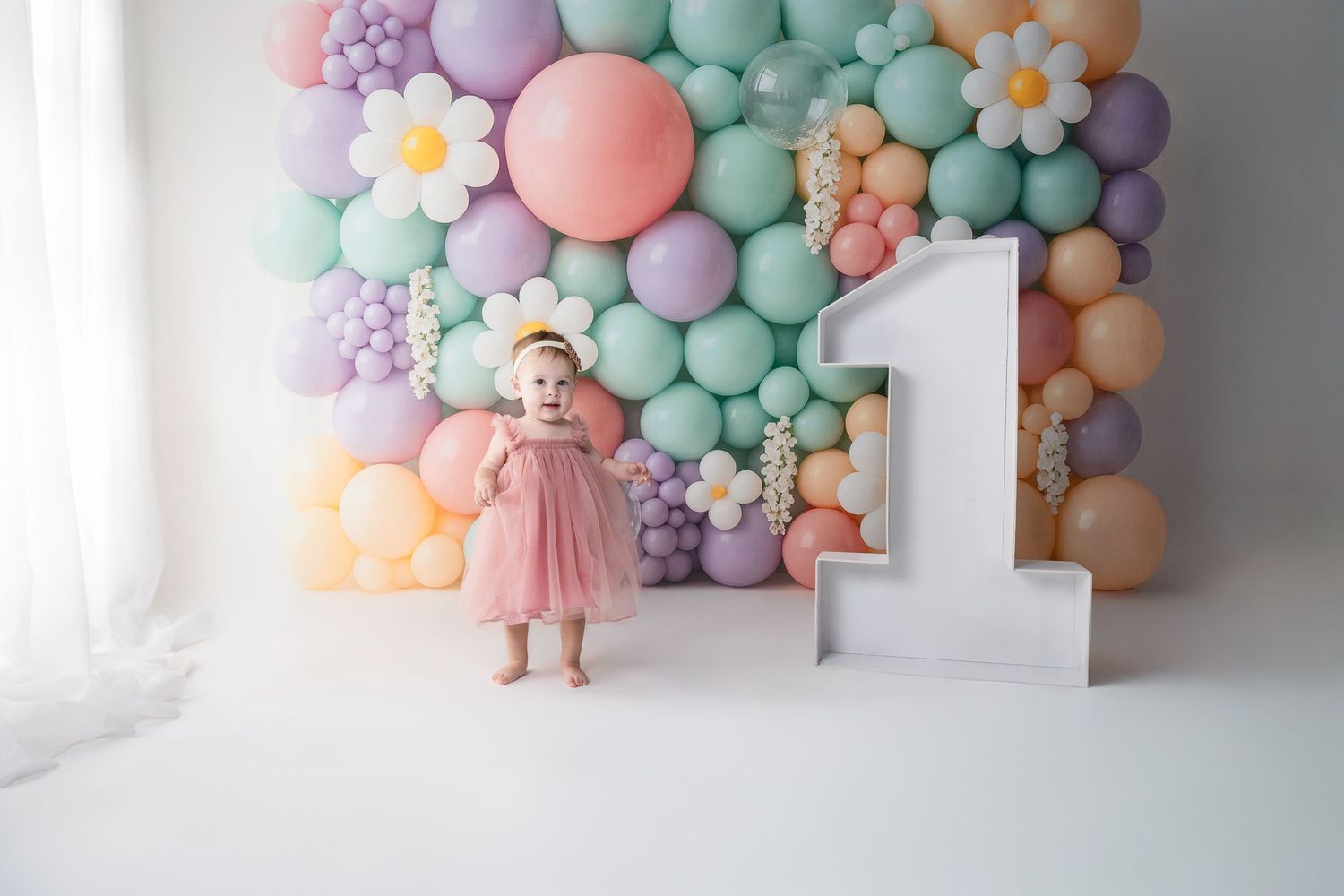 Pastel balloon arch with soft colors and floral accents against white wall.