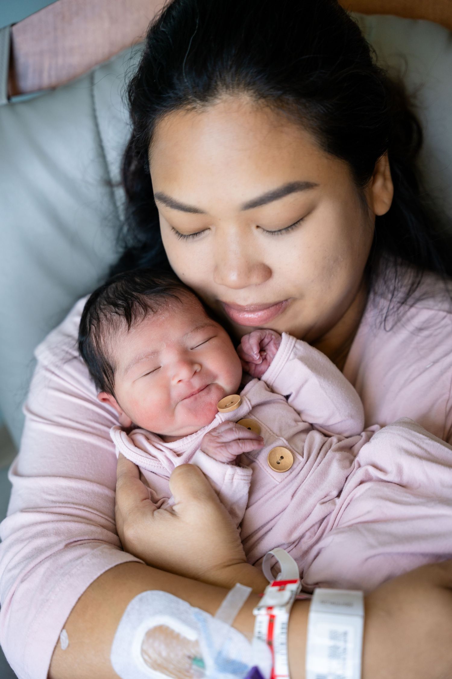 A new mother in a pink top wearing a hospital wristband gently cradles her sleeping newborn baby, who is wrapped in a soft pink outfit.