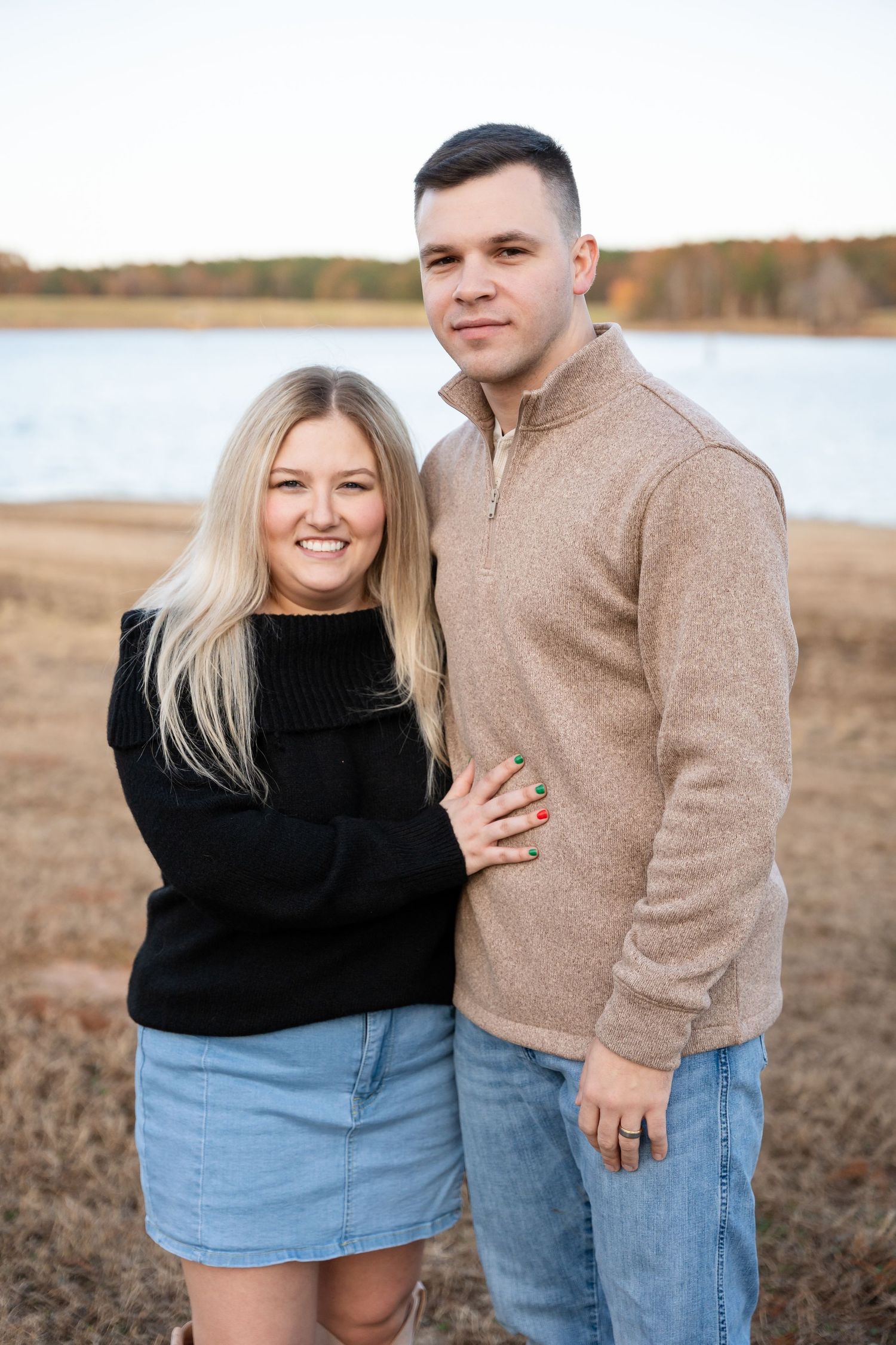 Smiling young couple embracing in front of a calm lake with autumn fields during an engagement photo session.