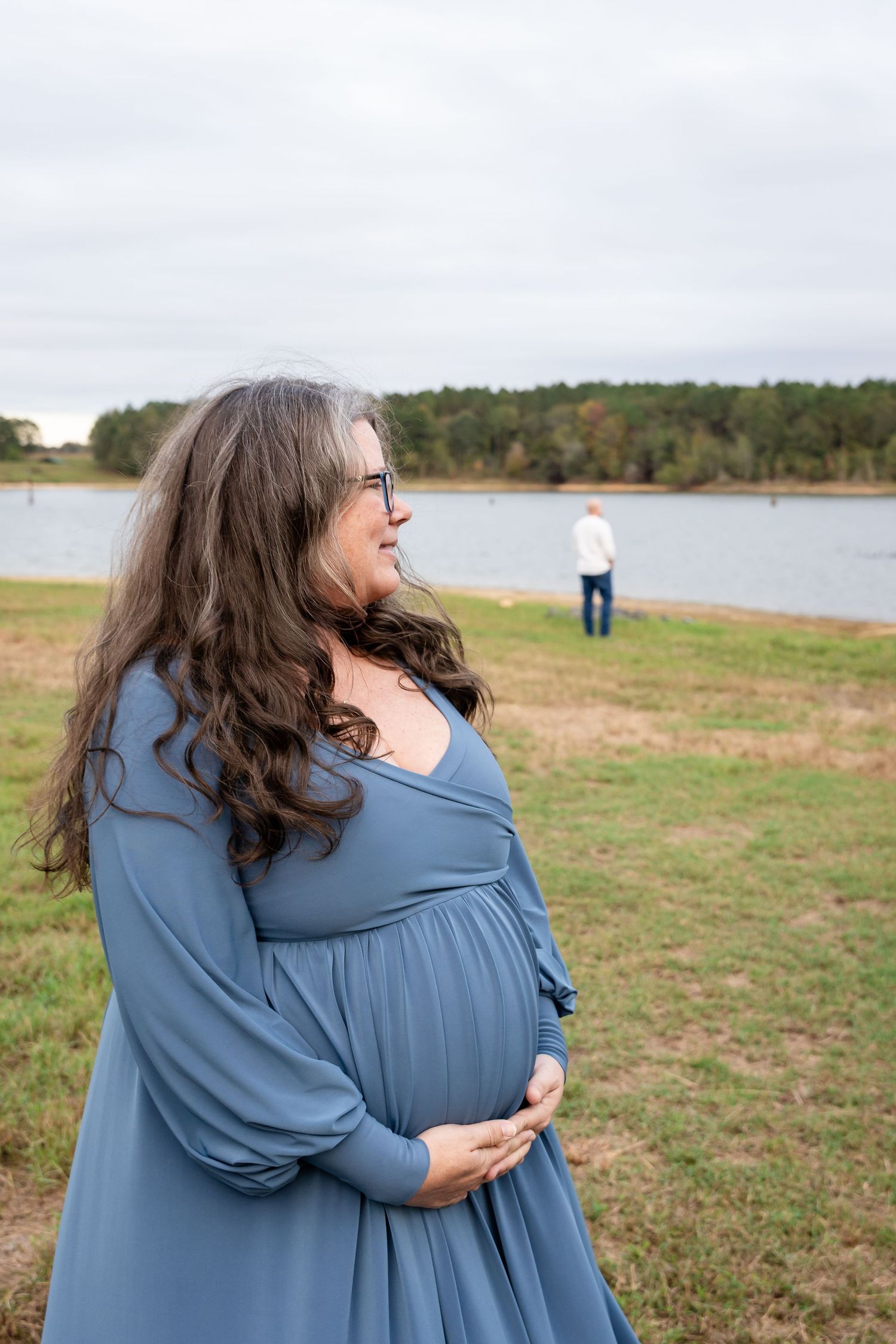 Two people embrace while standing in a scenic lakeside setting with trees in background.