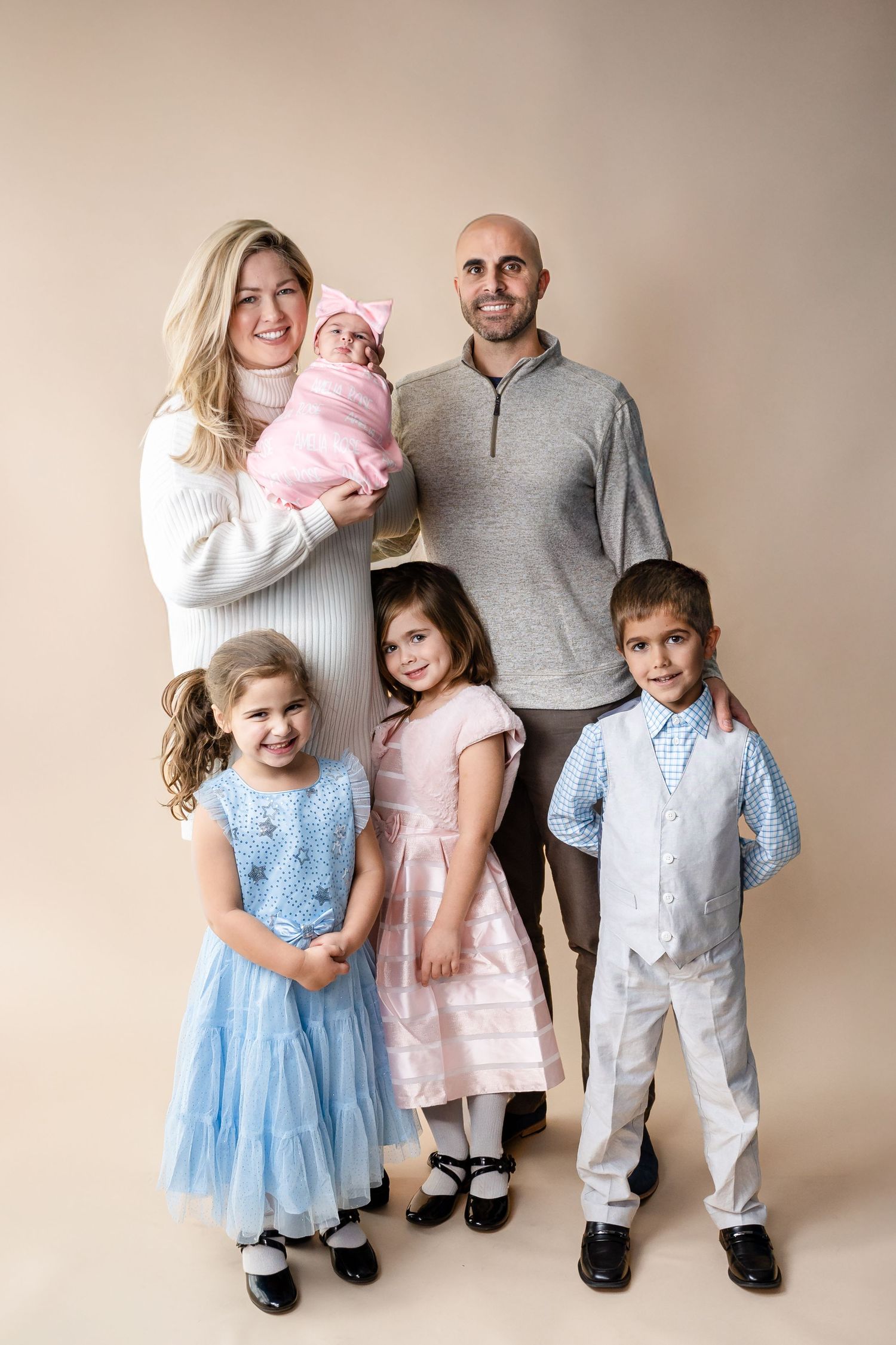 Family portrait with children in coordinating blue and pink formal outfits against beige background.