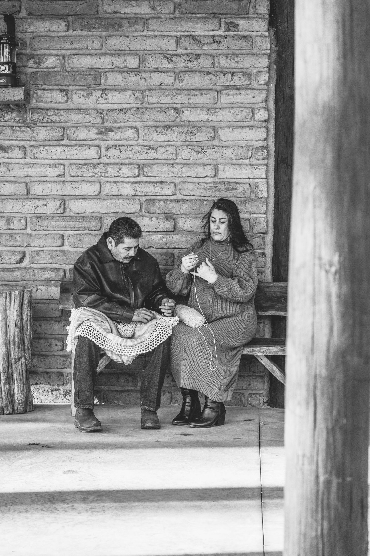 A black and white image of two people knitting together on a porch bench.