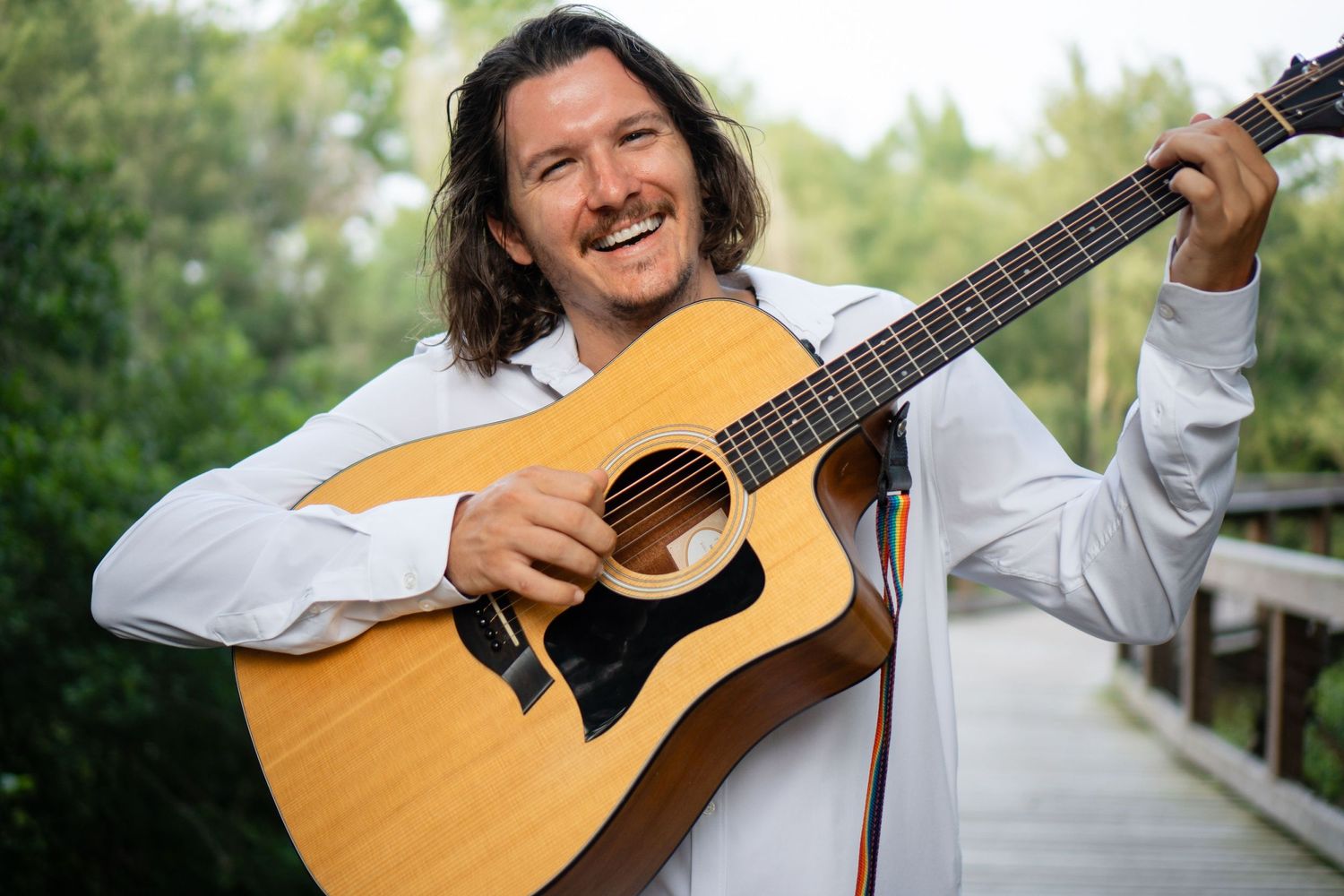 Musician playing acoustic guitar and smiling while standing outdoors in white shirt.