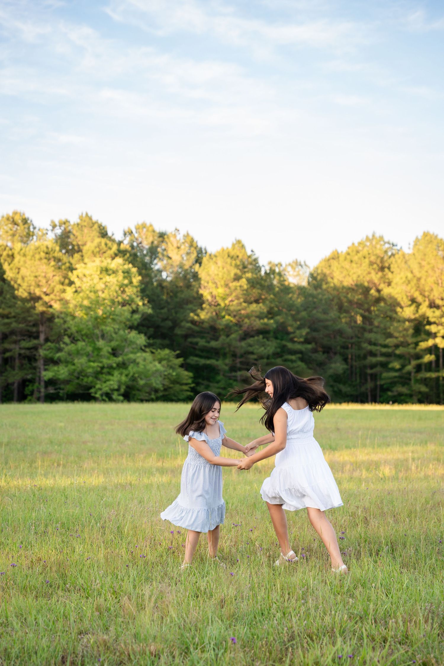 Two girls in white dresses hold hands and spin joyfully in a sunlit green meadow with tall trees in the background.