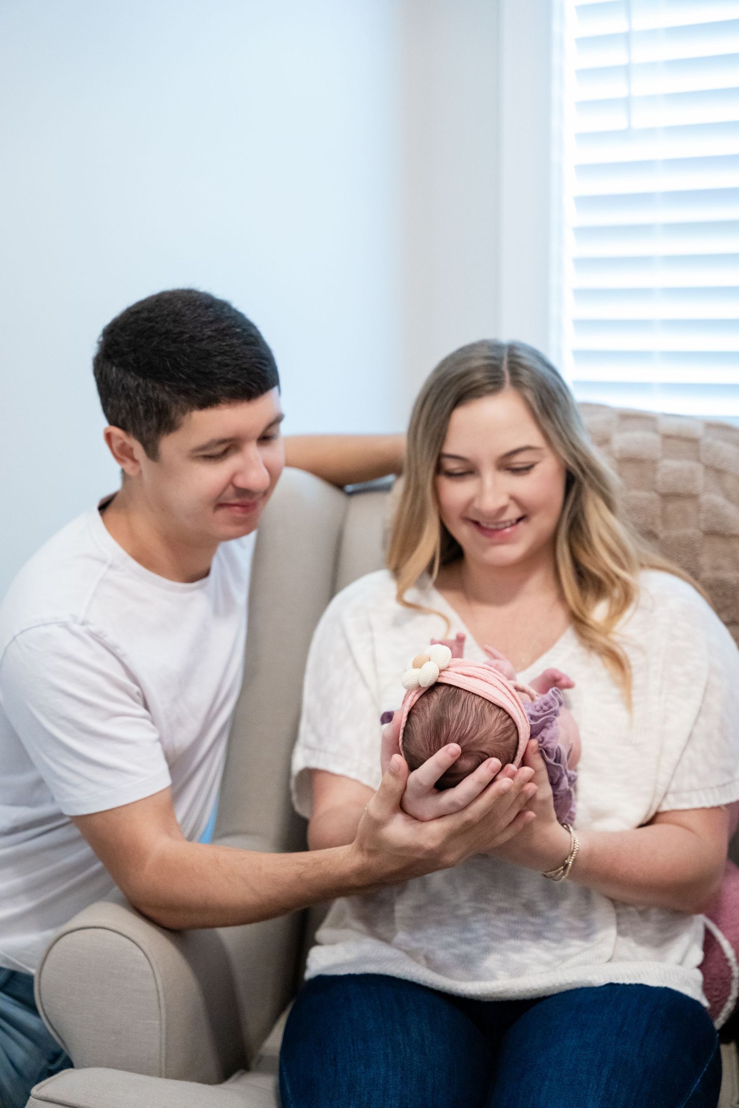 A couple smiles while holding their newborn baby wrapped in pink on a couch indoors.