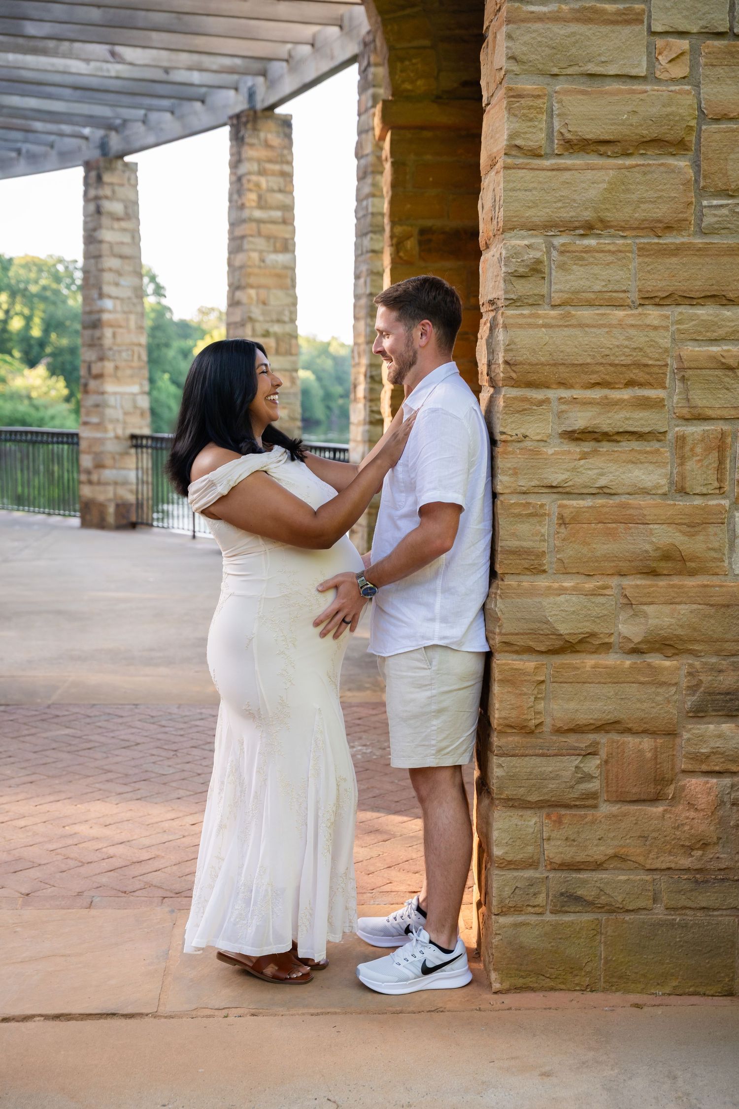 Expecting couple in a white dress and casual shirt embracing beside a stone pillar during a sunny outdoor maternity photo session.