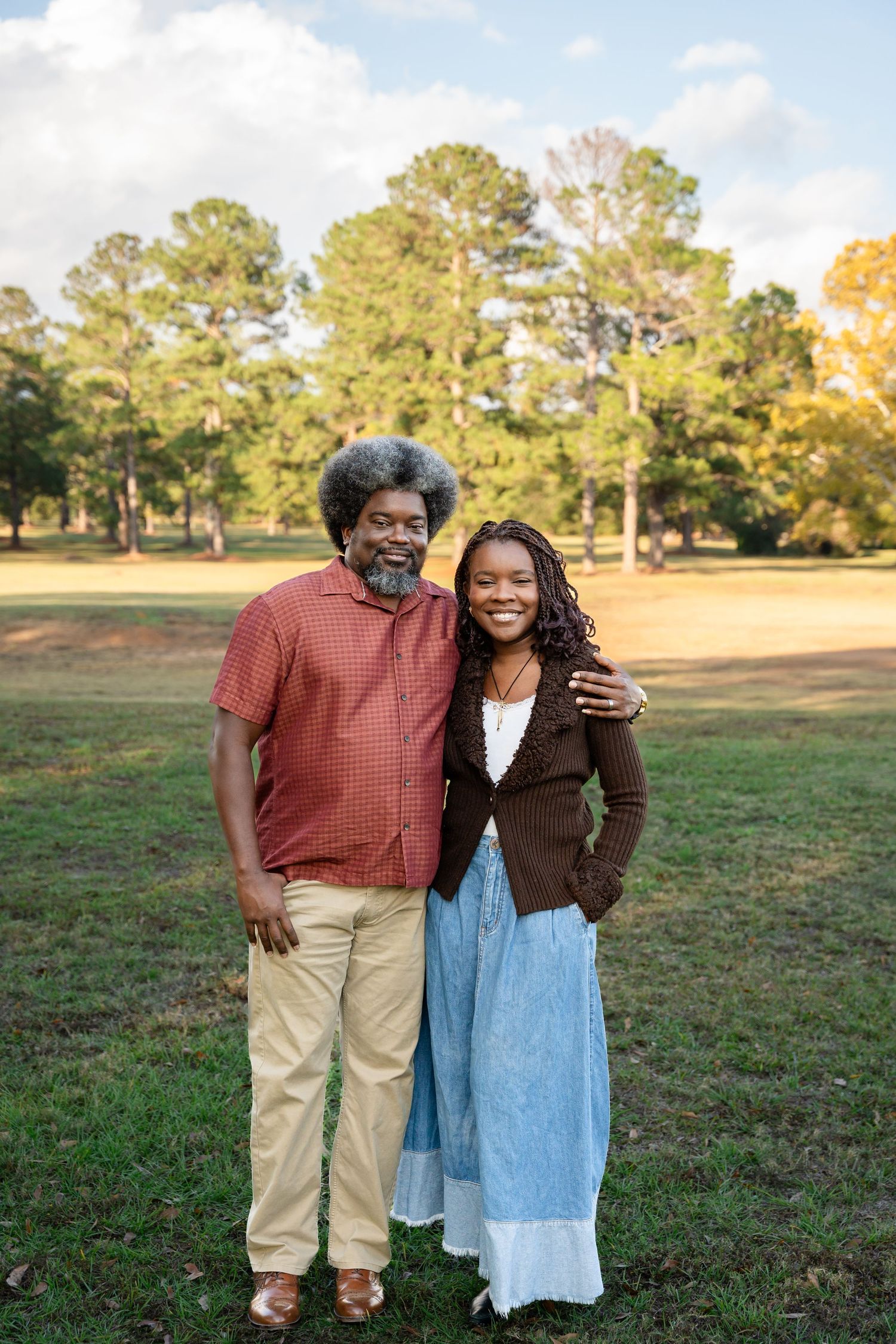 Two people embracing while standing in a sunny field with pine trees and autumn foliage.