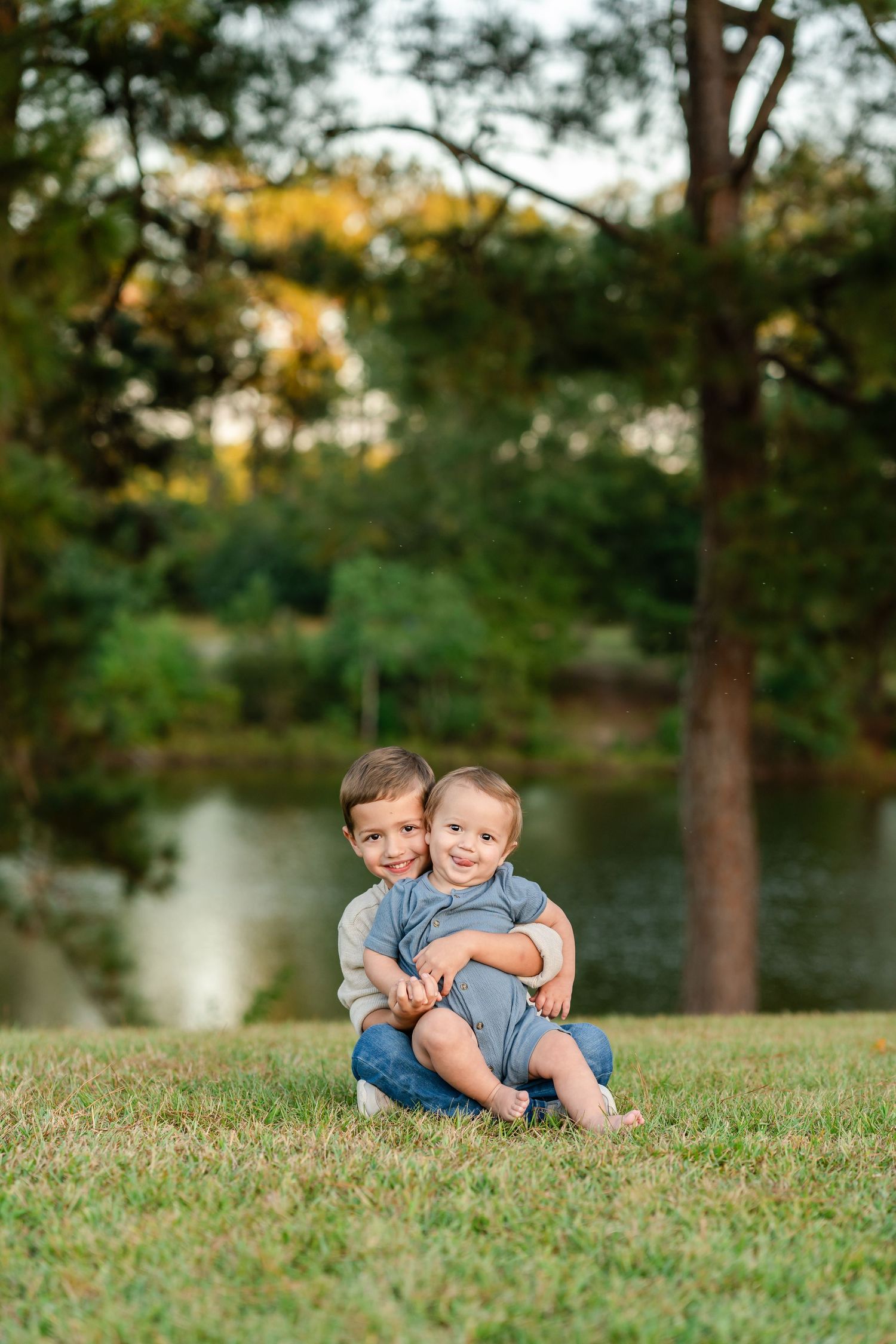 Two children sit together on grassy lawn near a tranquil pond at sunset with trees in background.
