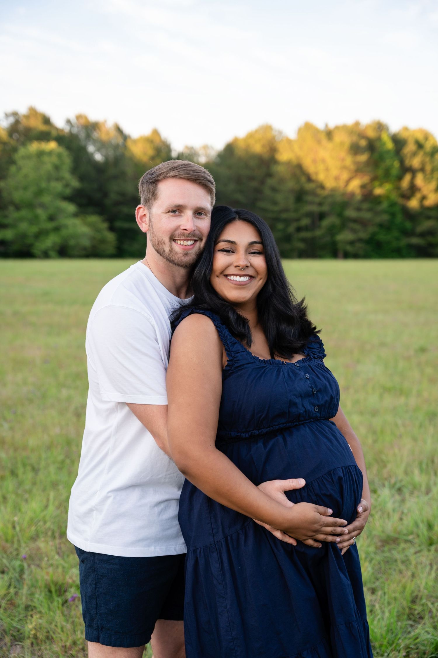 Smiling pregnant couple embracing in a green field at sunset, woman in navy dress.