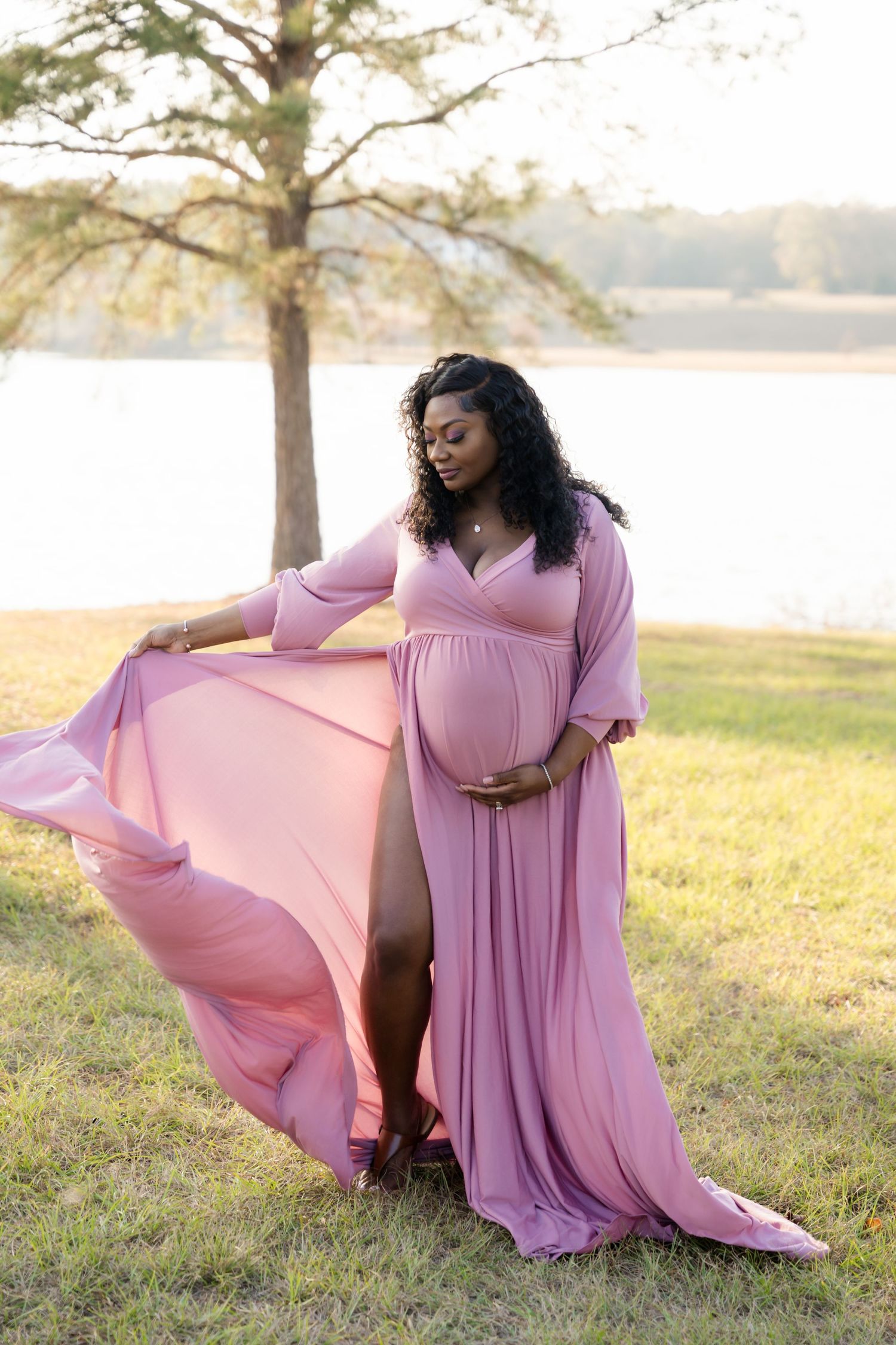 Pregnant woman in flowing pink maternity gown poses gracefully outdoors near a lake at golden hour.