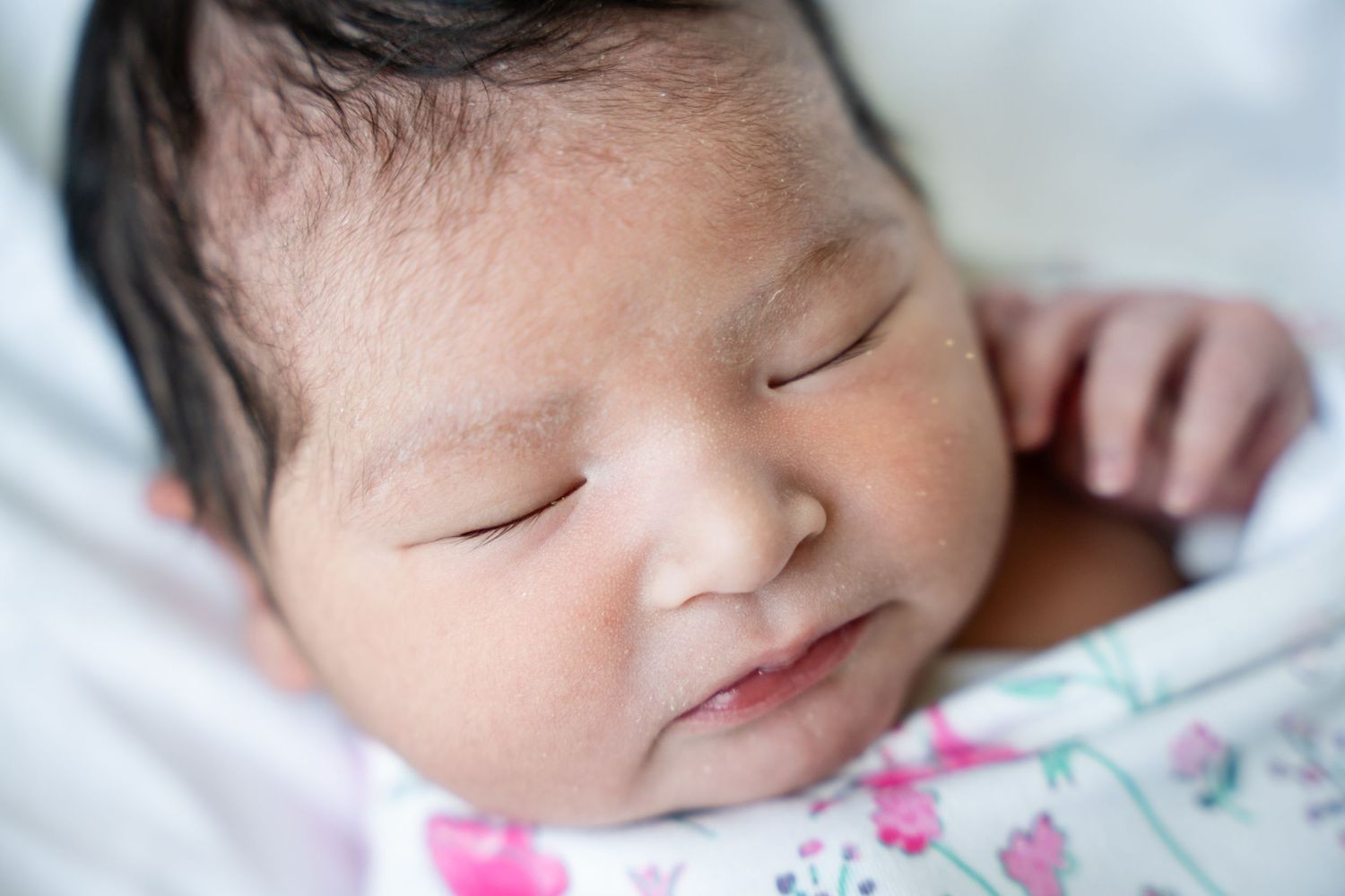 Close-up of a sleeping newborn baby with dark hair, wrapped in a soft blanket with a pink floral print.