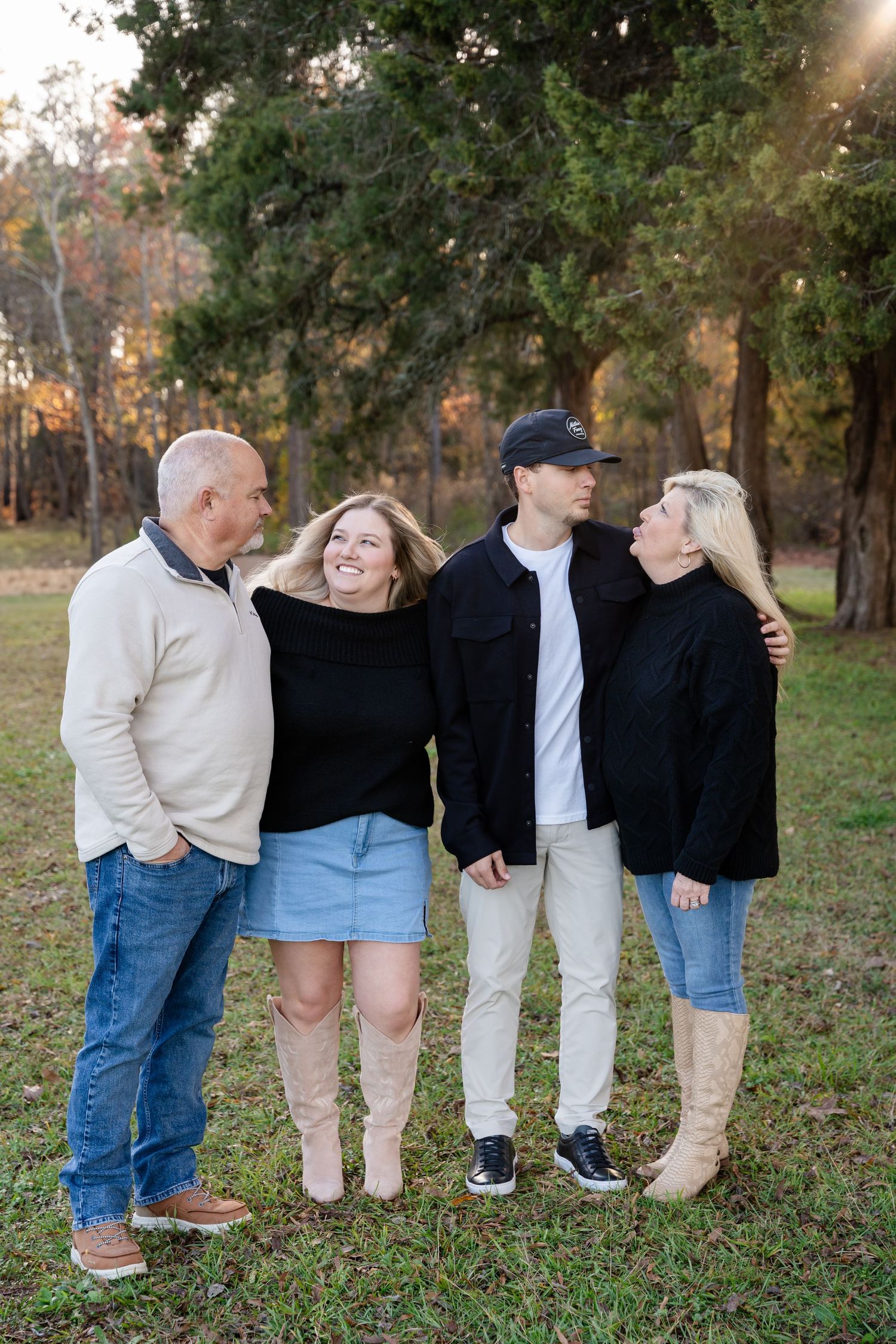 Family of four standing together on a grassy lawn among tall trees during a golden hour outdoor portrait session.