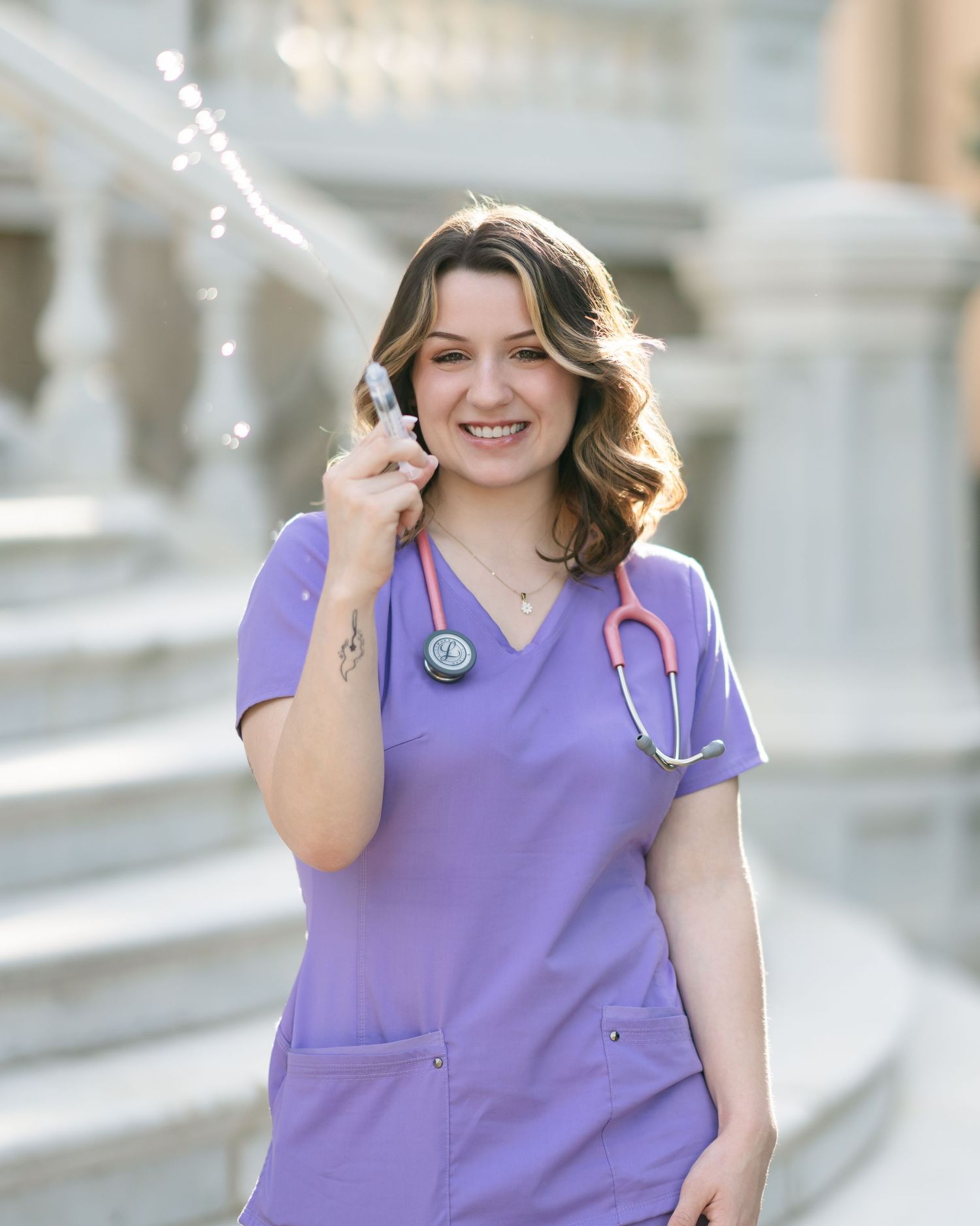 Medical professional in purple scrubs with stethoscope posing outdoors by white architectural features.