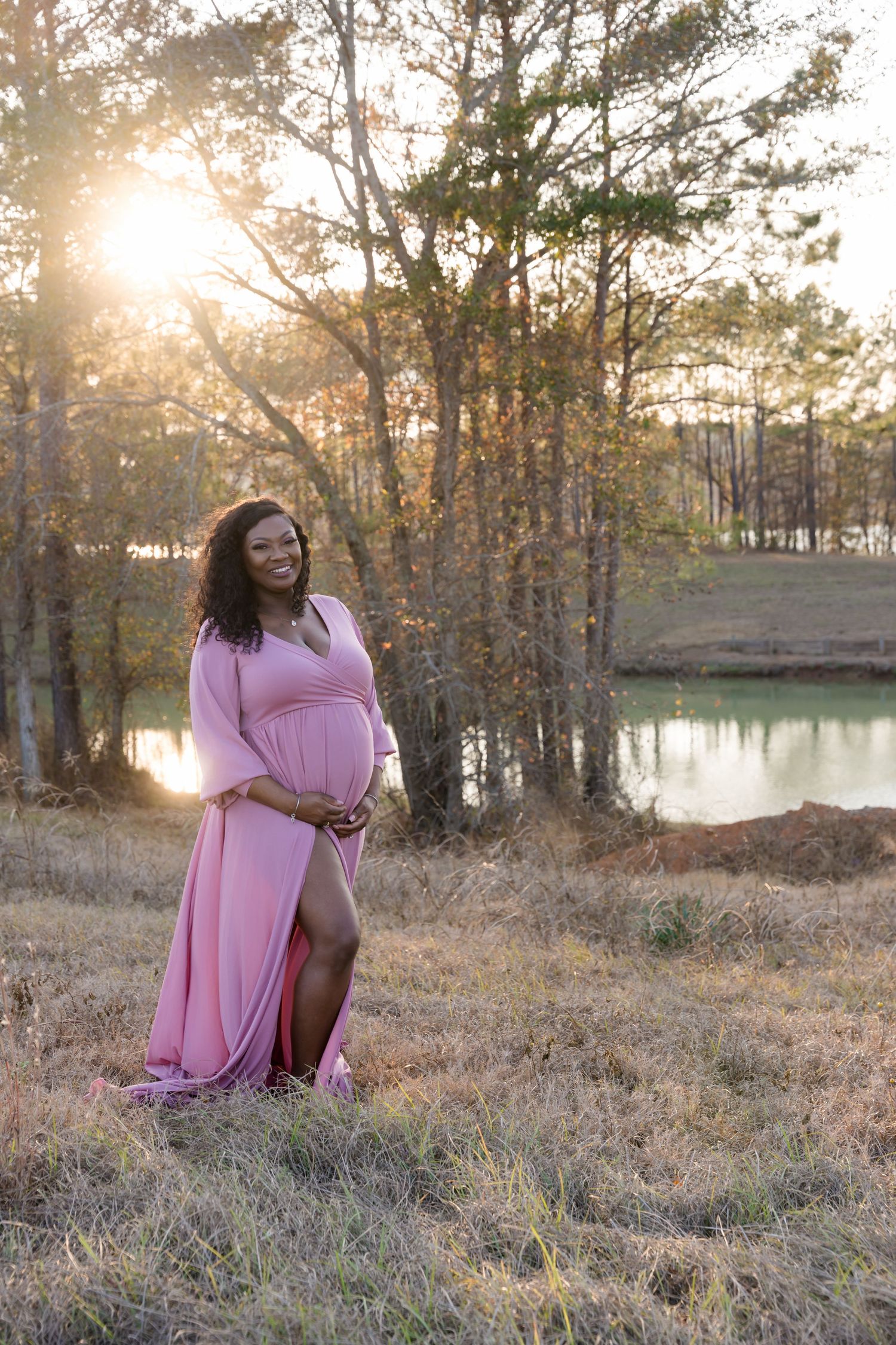 Smiling pregnant woman in pink maternity gown poses outdoors at golden hour near a serene lake surrounded by trees.