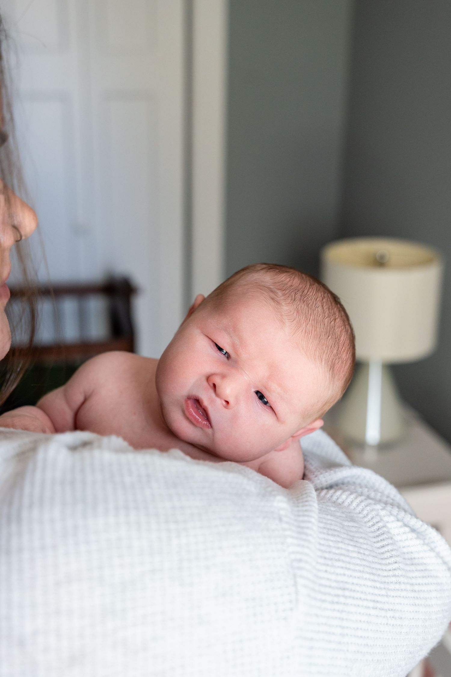 Newborn baby lying on a white blanket, alert and awake, held by a parent in a softly lit nursery room.