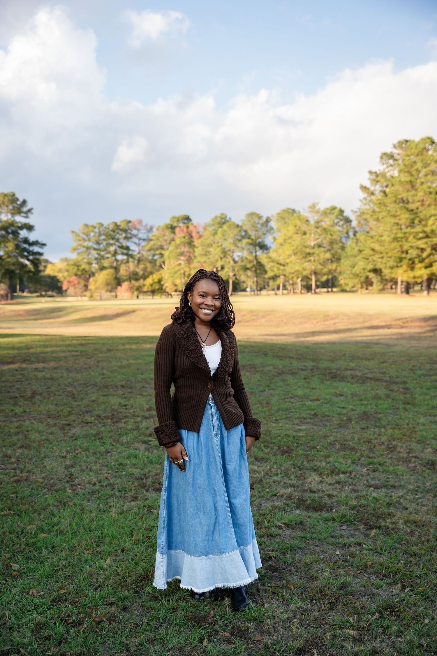 Person in dark cardigan and blue dress poses expressively in grassy field.