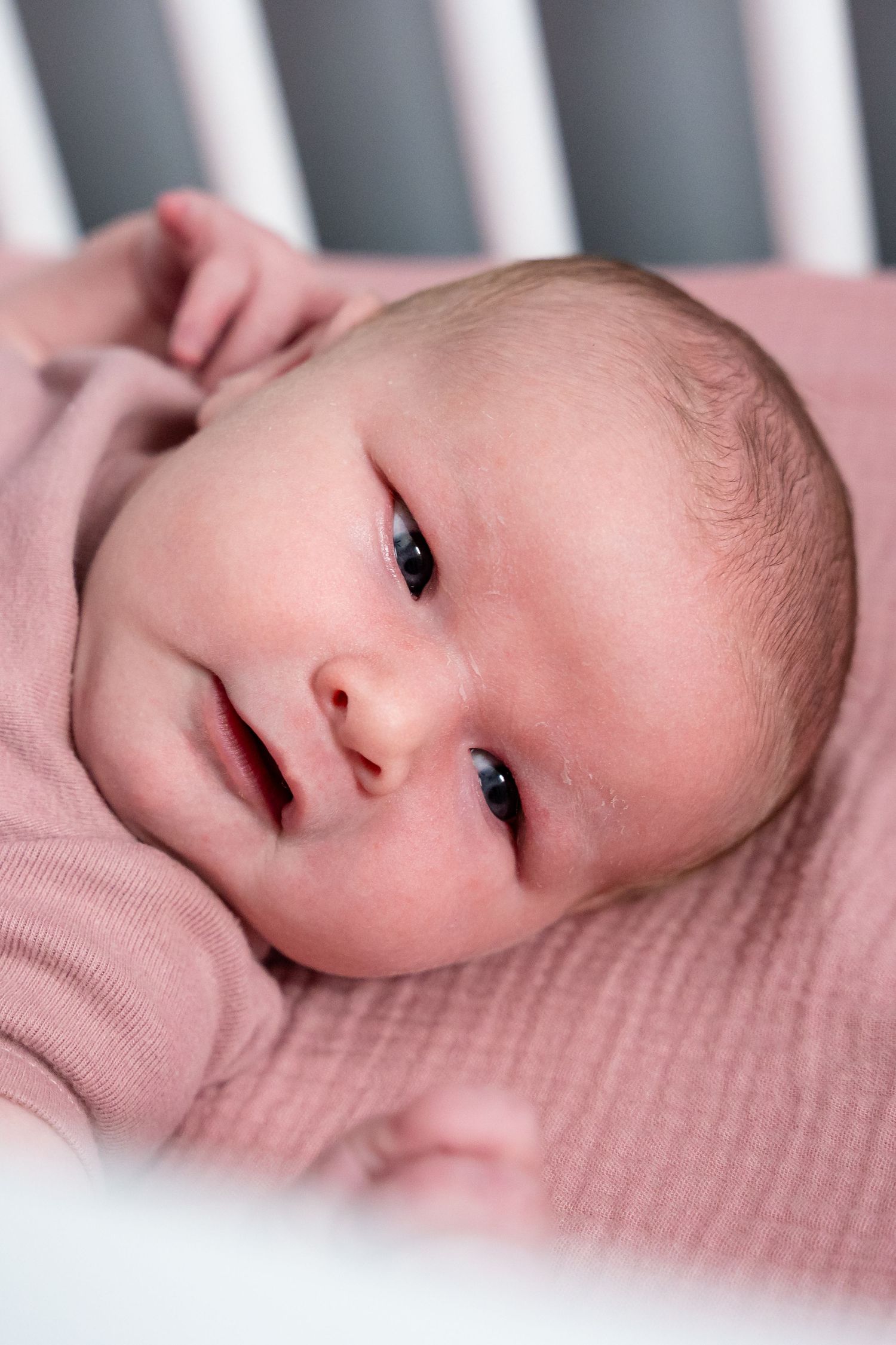 Newborn baby in pink outfit lying awake in a white crib, with bright eyes and tiny hands visible.