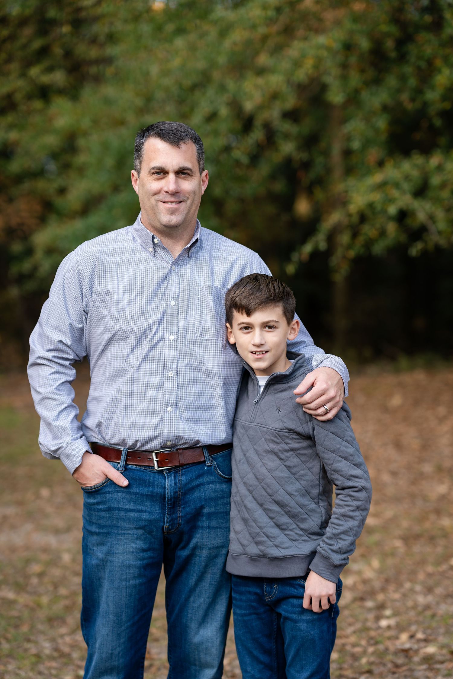 Father and son stand together outdoors in coordinating gray shirts against a wooded backdrop.