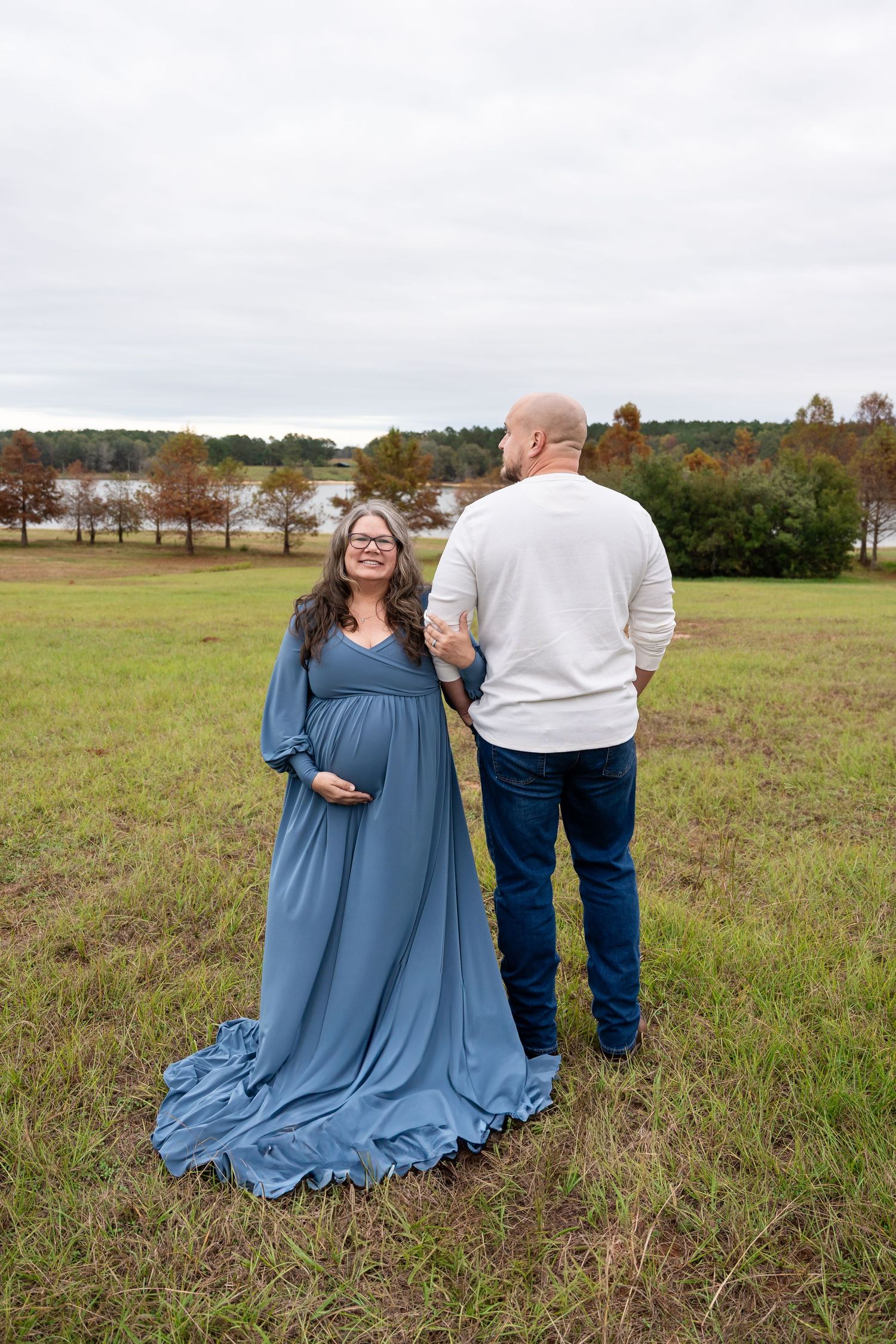 Outdoor maternity photos in a grassy field with fall trees and water in background.
