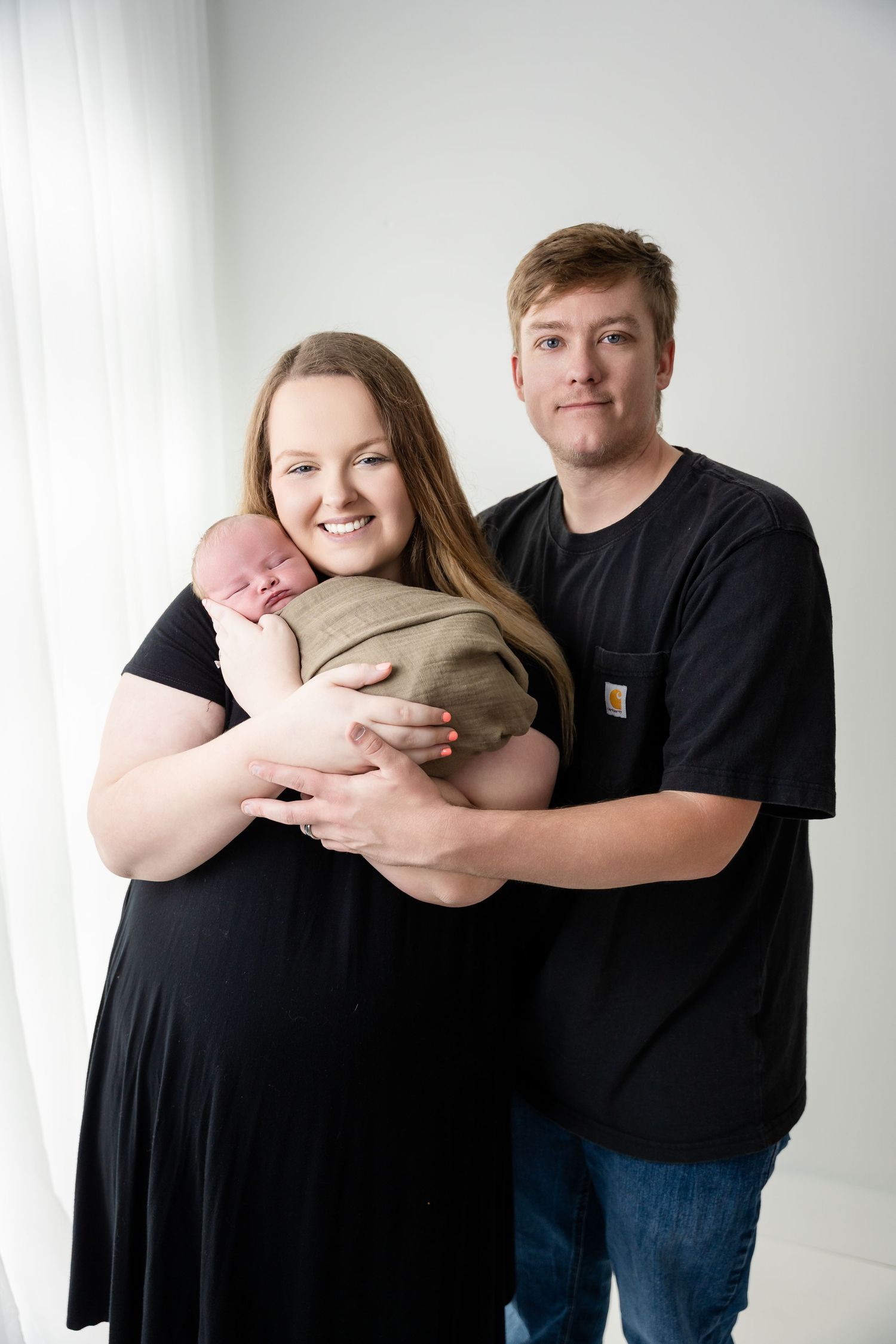 A couple in black clothing share tender moments with their newborn baby in a white studio setting.