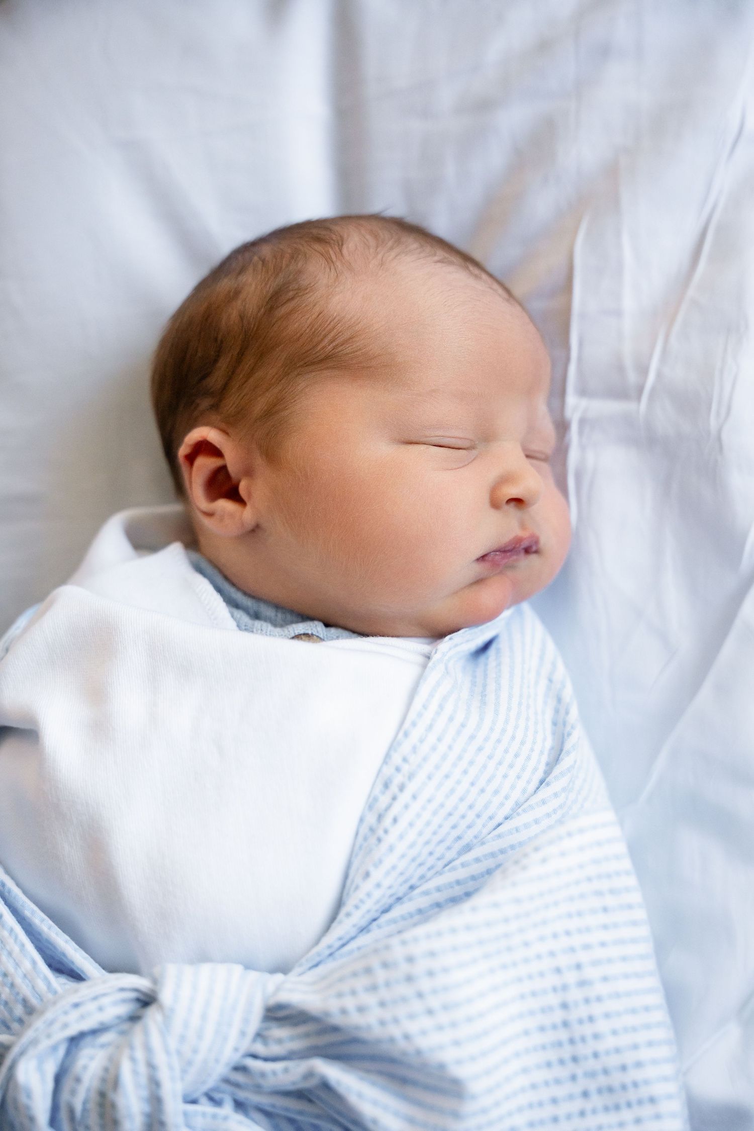Baby wearing light blue clothing sleeps soundly on white sheets.