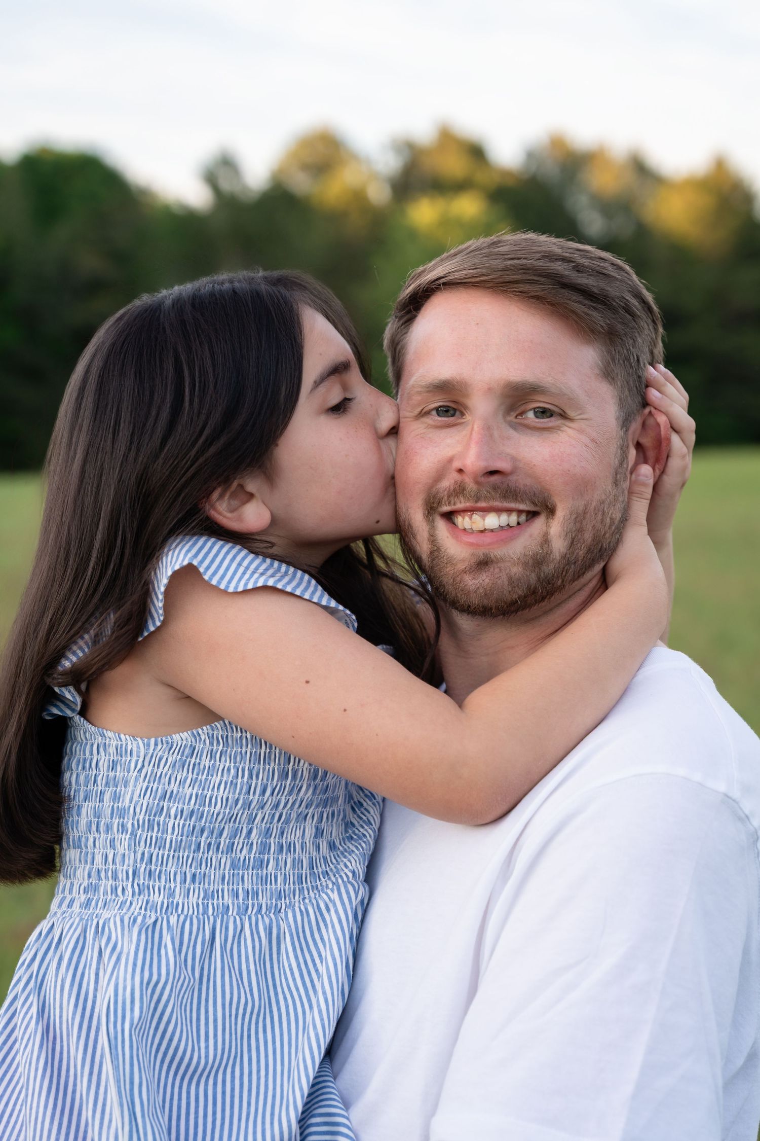 A young girl in a blue dress kisses a smiling bearded man on the cheek outdoors.