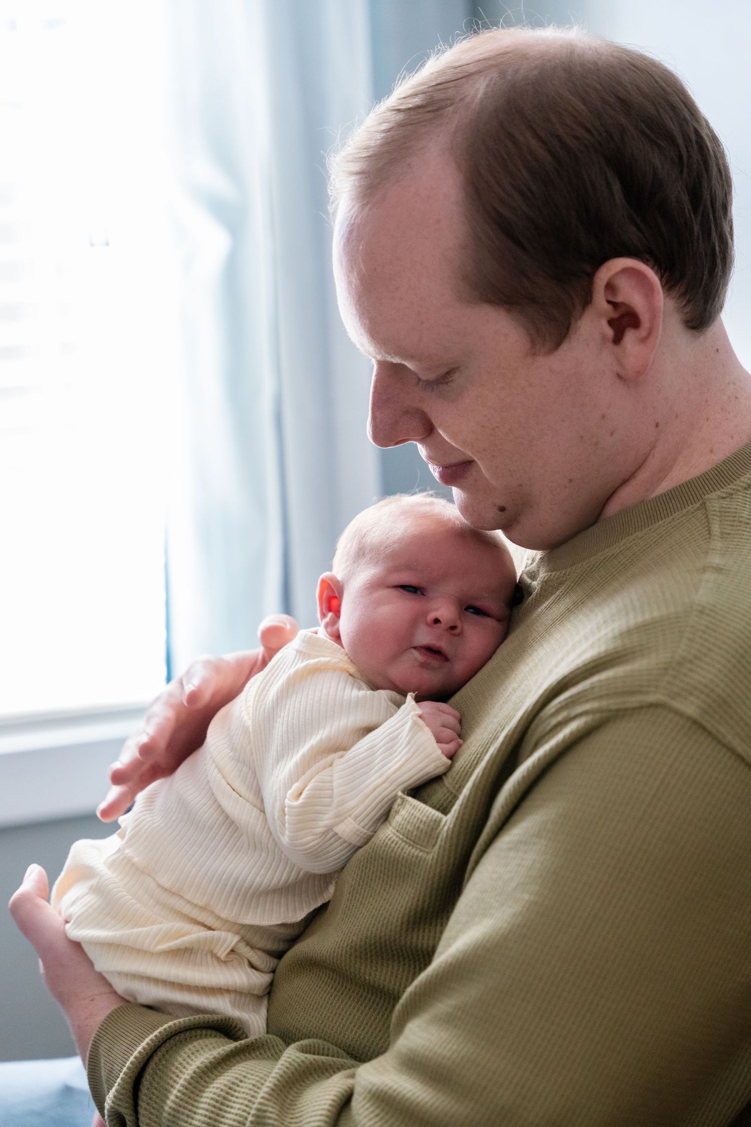 A father tenderly holds his newborn baby against his chest near a bright window.