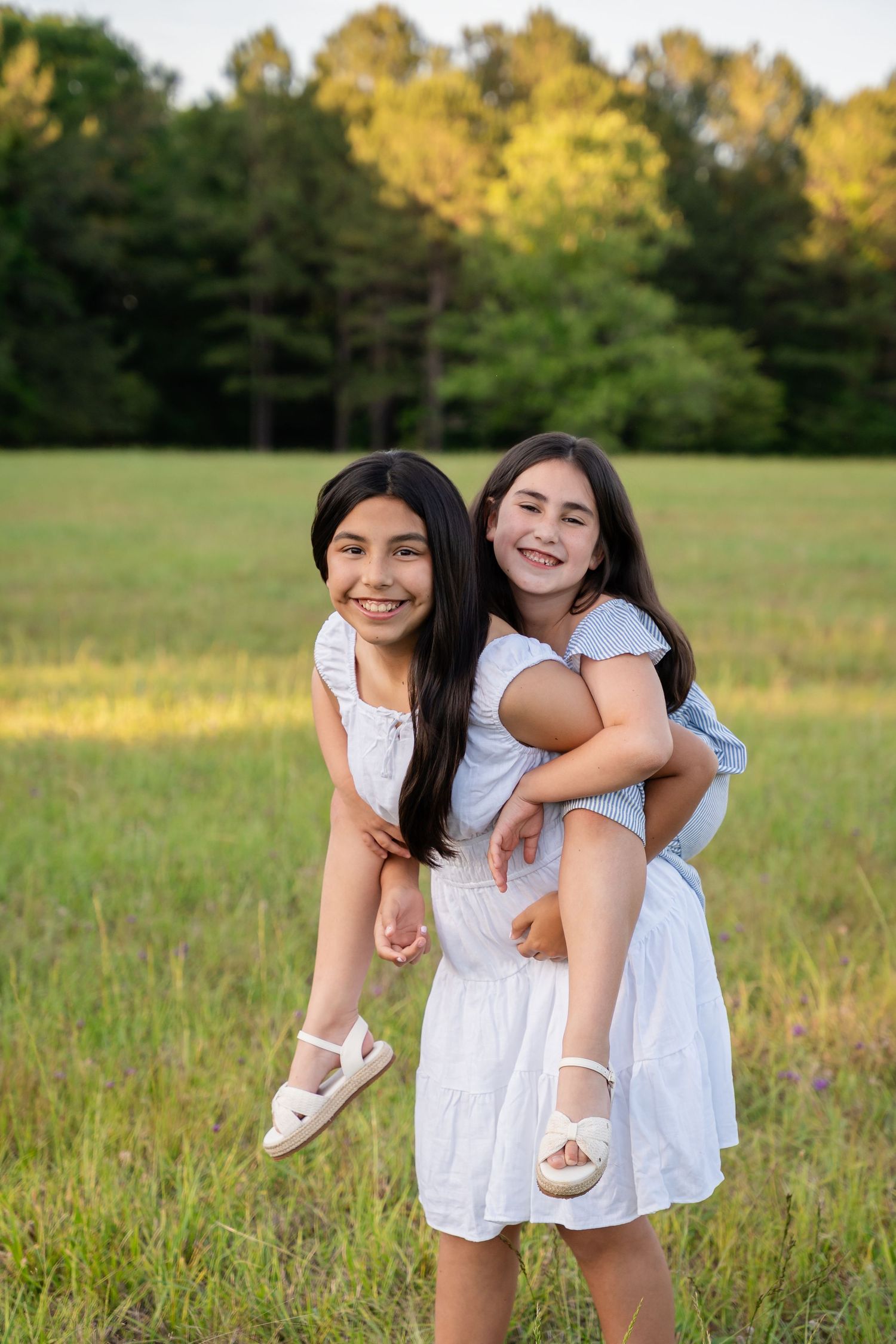 Two happy girls in white dresses and sandals enjoy a piggyback ride in a sunlit grassy field with trees in the background.