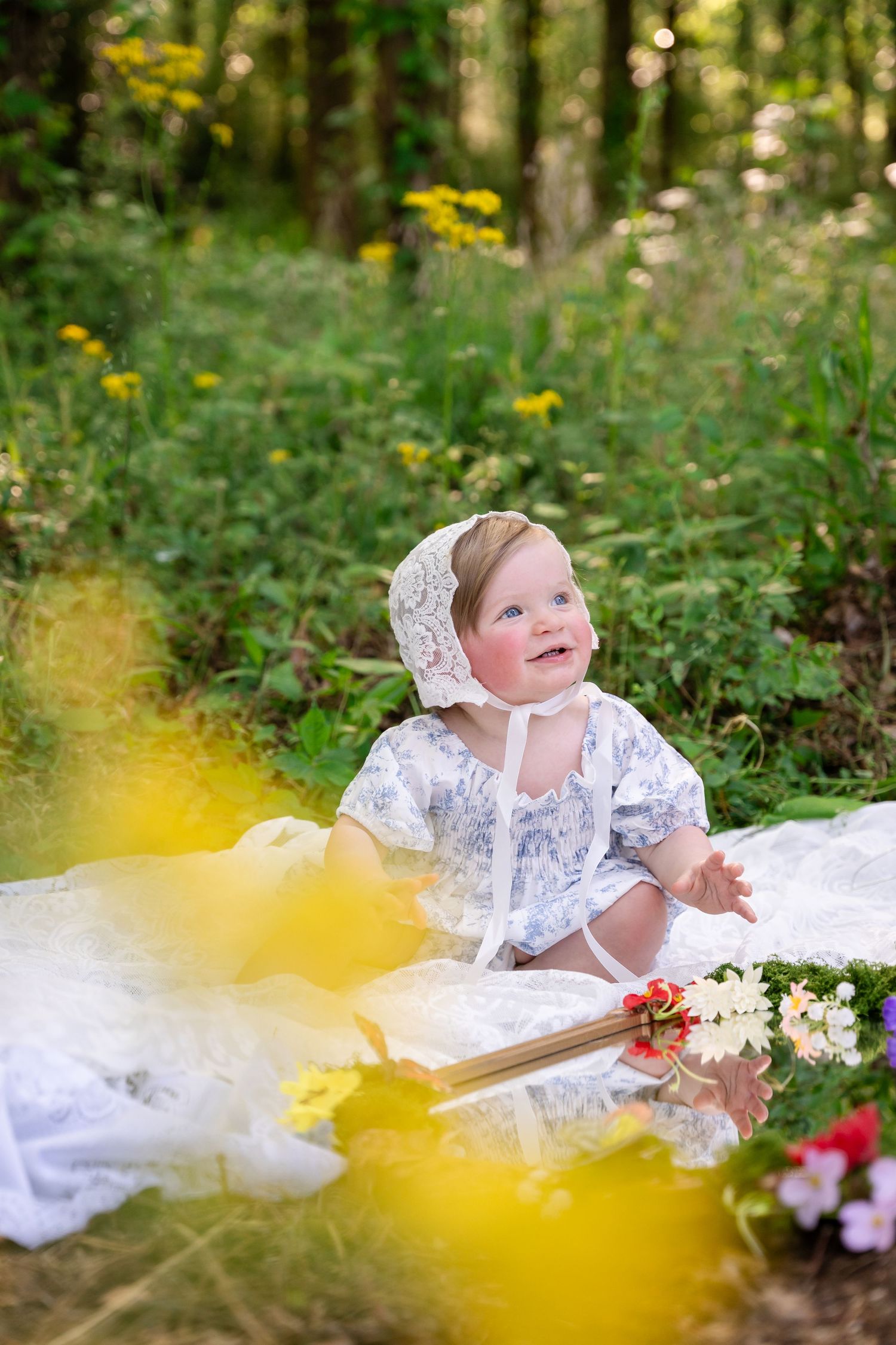 Baby girl in lace bonnet and floral dress sits on white blanket surrounded by wildflowers in a sunny forest setting.