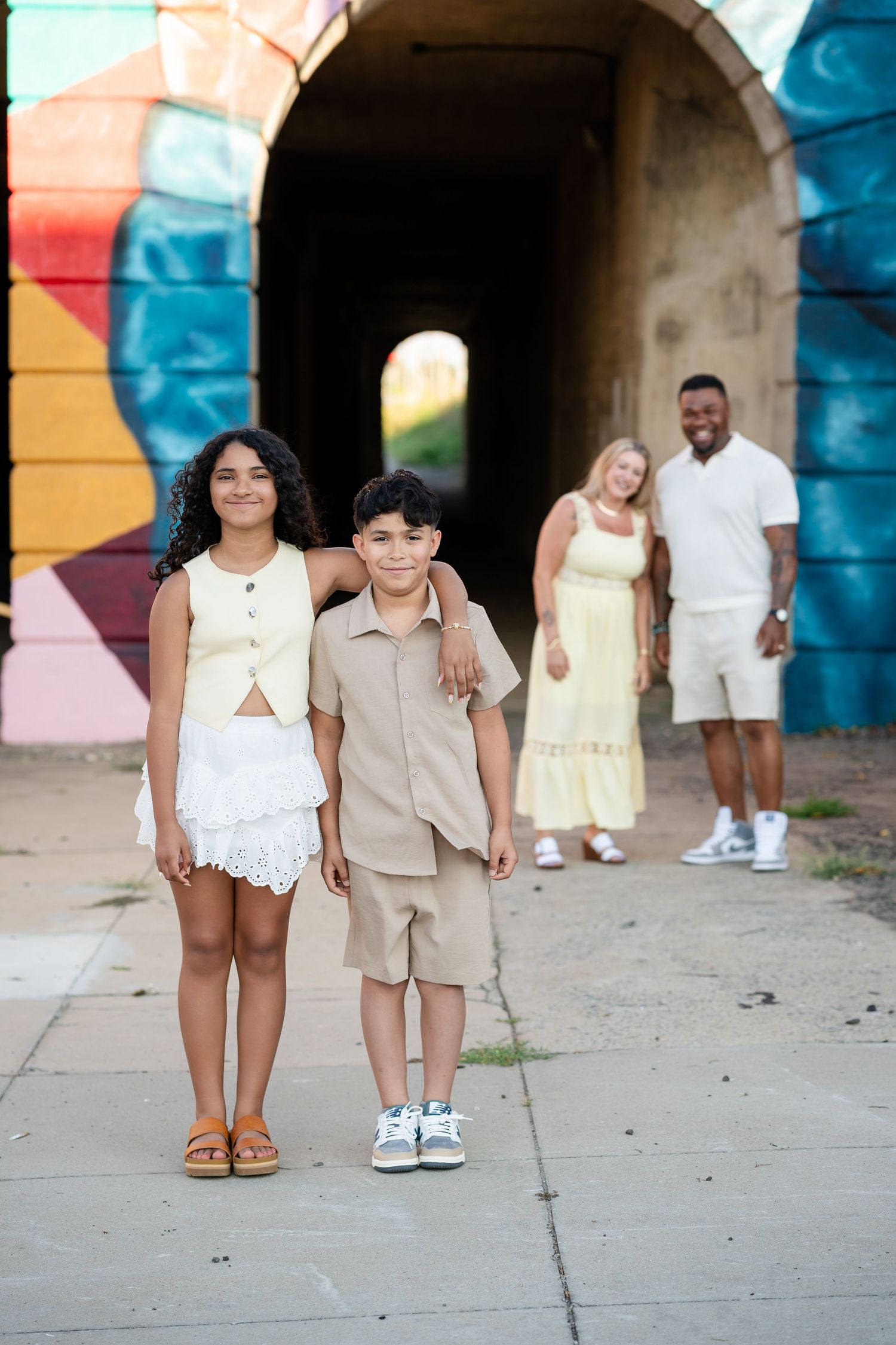Two children pose in front of a colorful mural tunnel while their parents stand behind them.