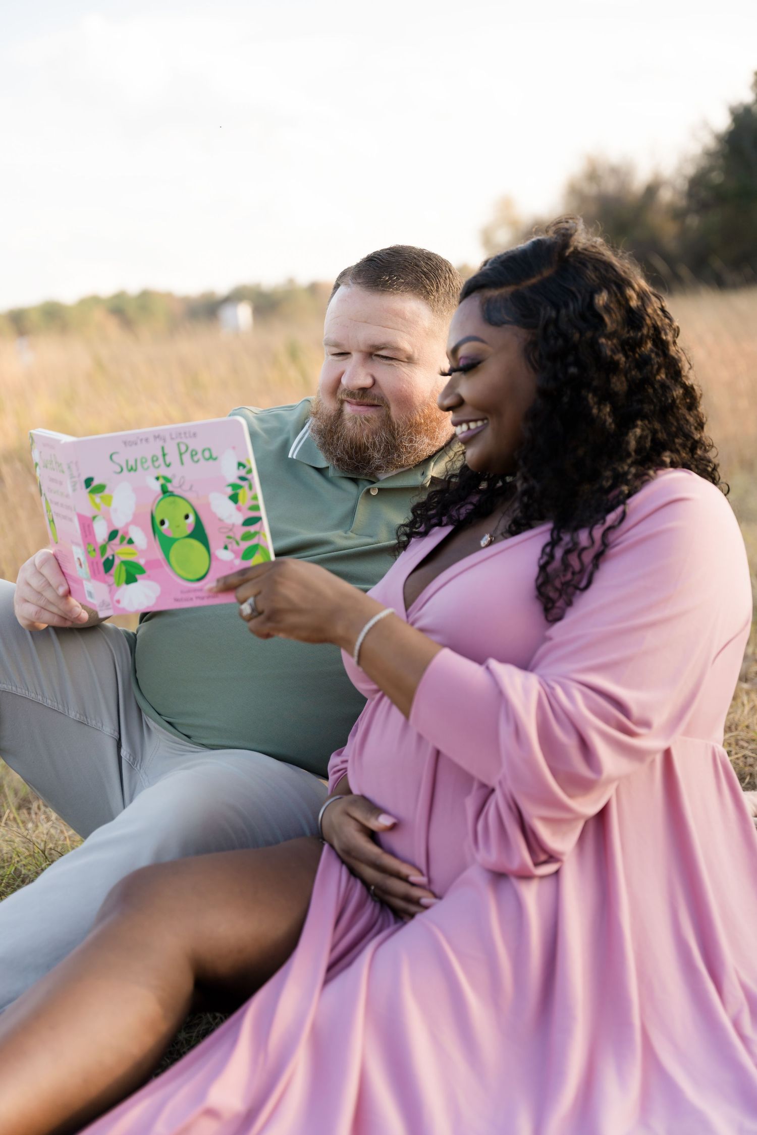 Pregnant woman in pink dress and man reading Sweet Pea book together outdoors in a golden field maternity photo.