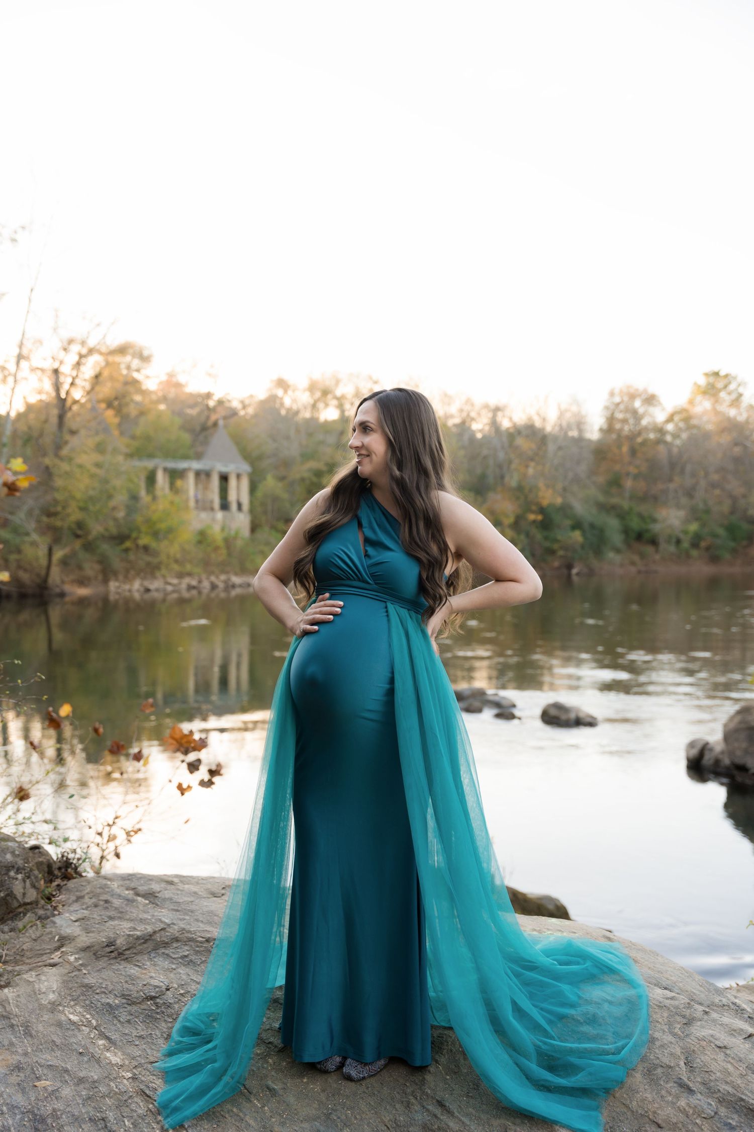 A person in a teal dress and flowing cape poses dramatically by a serene lake during autumn.