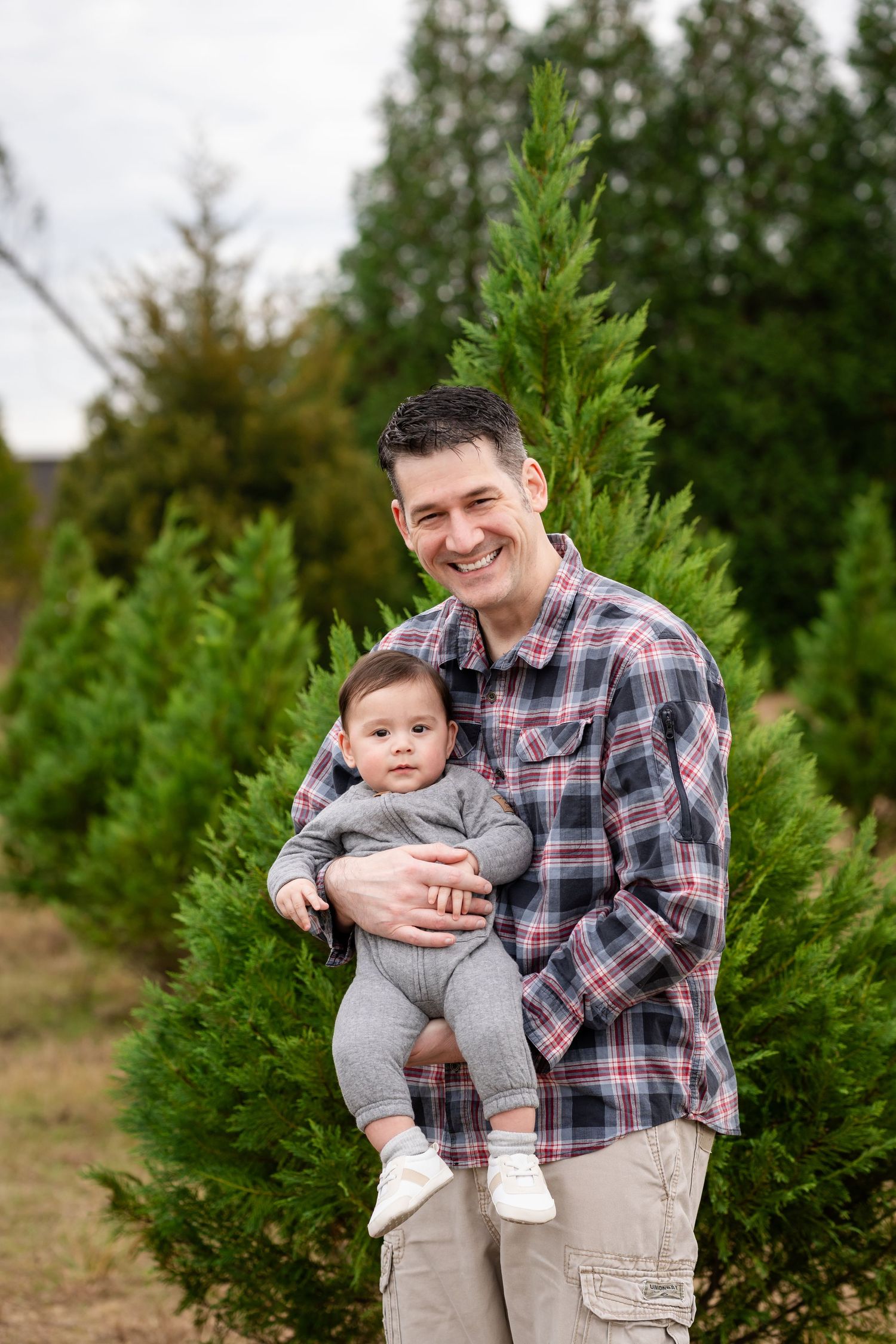 Parent and child sharing joyful expressions during outdoor portrait session among evergreen trees.