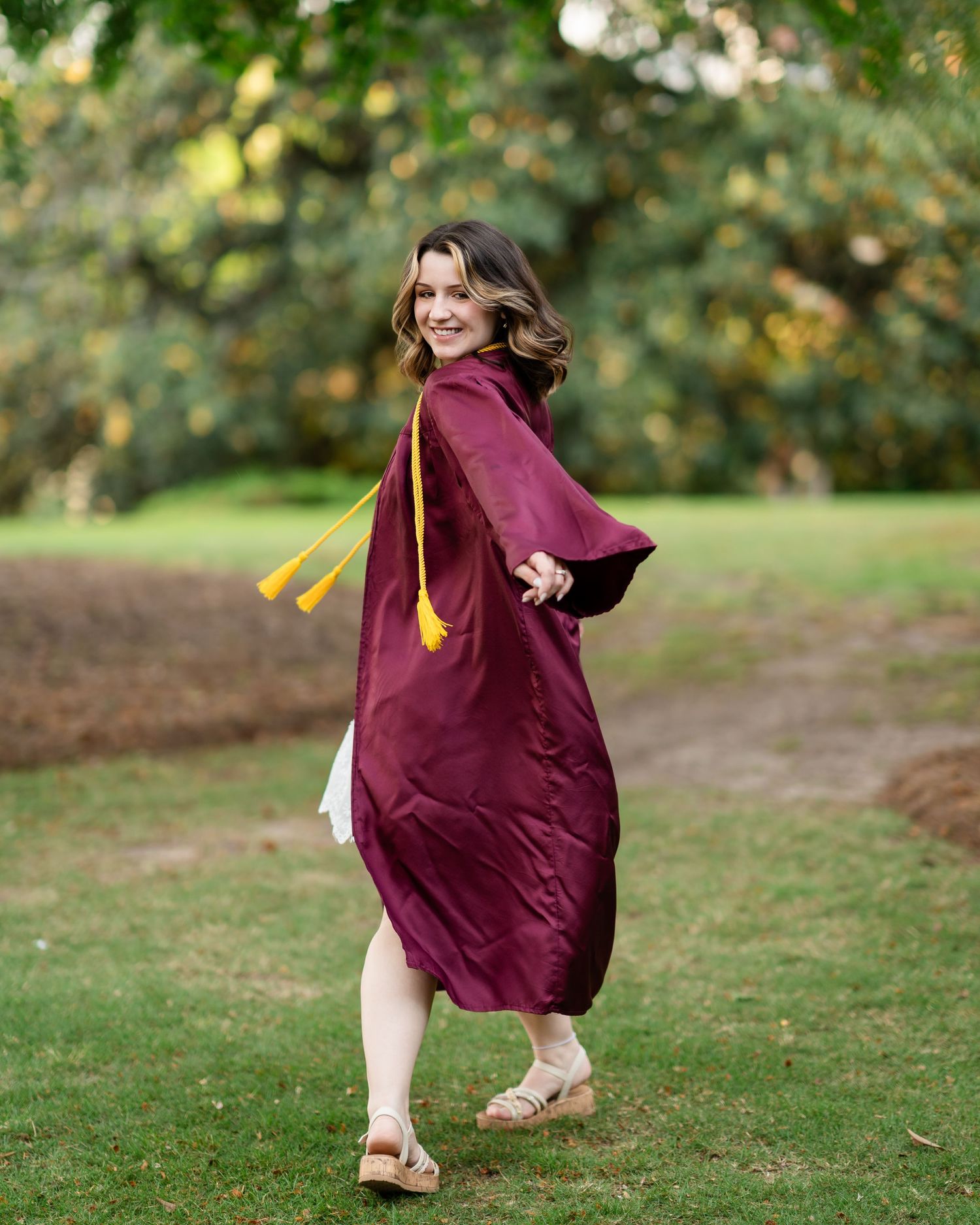 Person in white dress and burgundy graduation gown twirling in a garden setting.