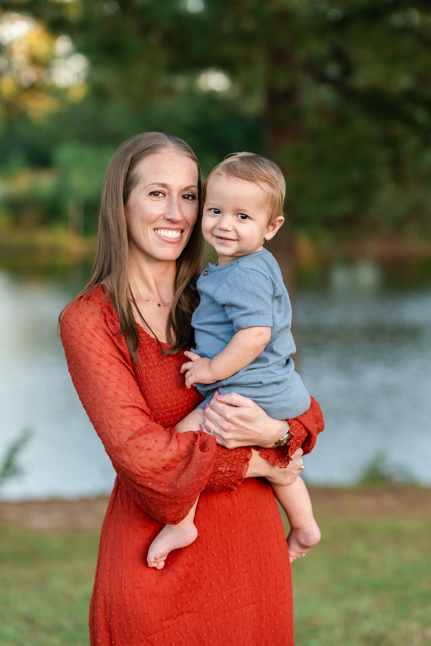 A smiling woman in a red dress holds a happy baby boy in a blue outfit outdoors near a pond.