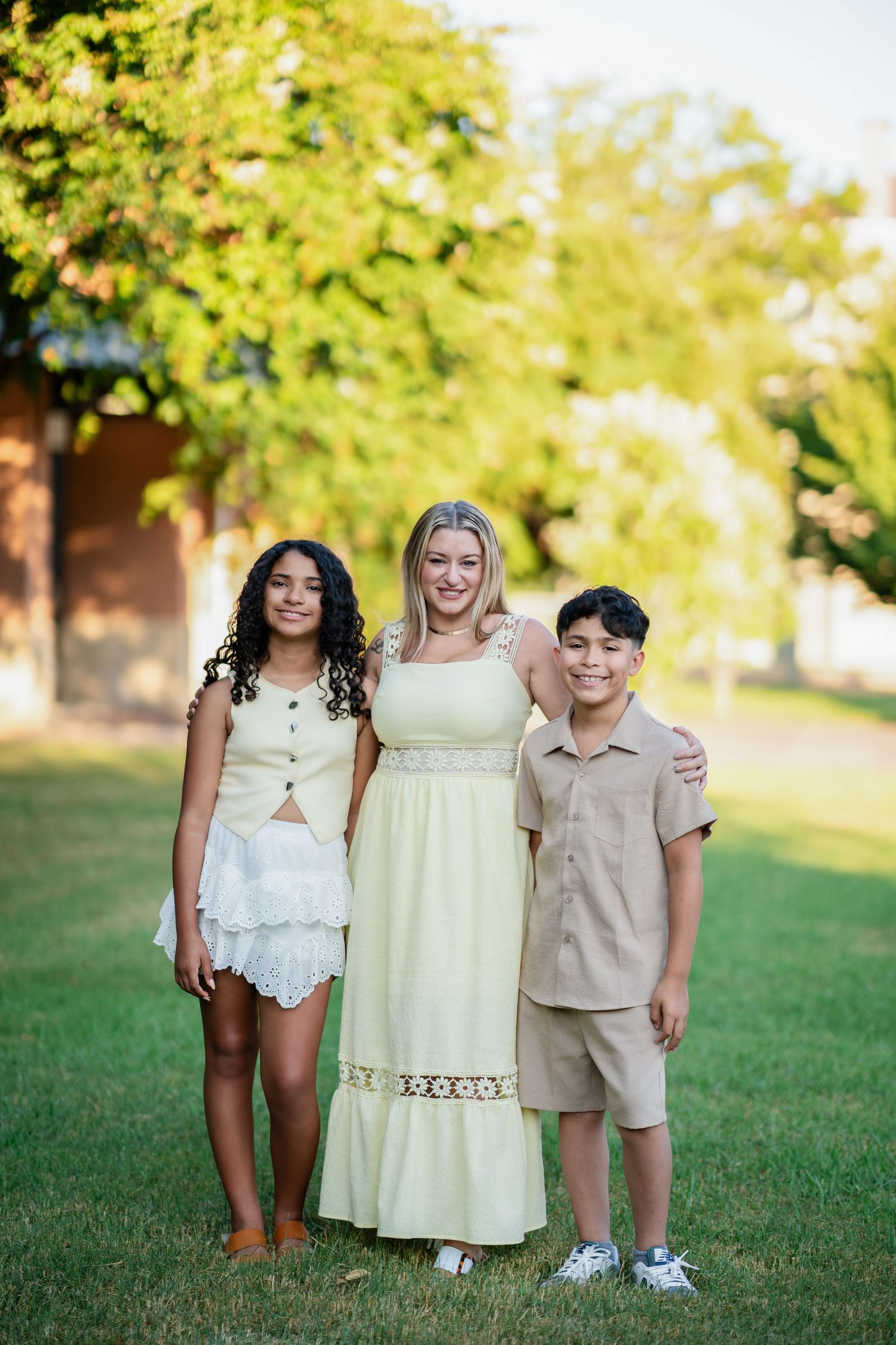 A woman in a yellow maxi dress poses outdoors with two children in a sunny green park.