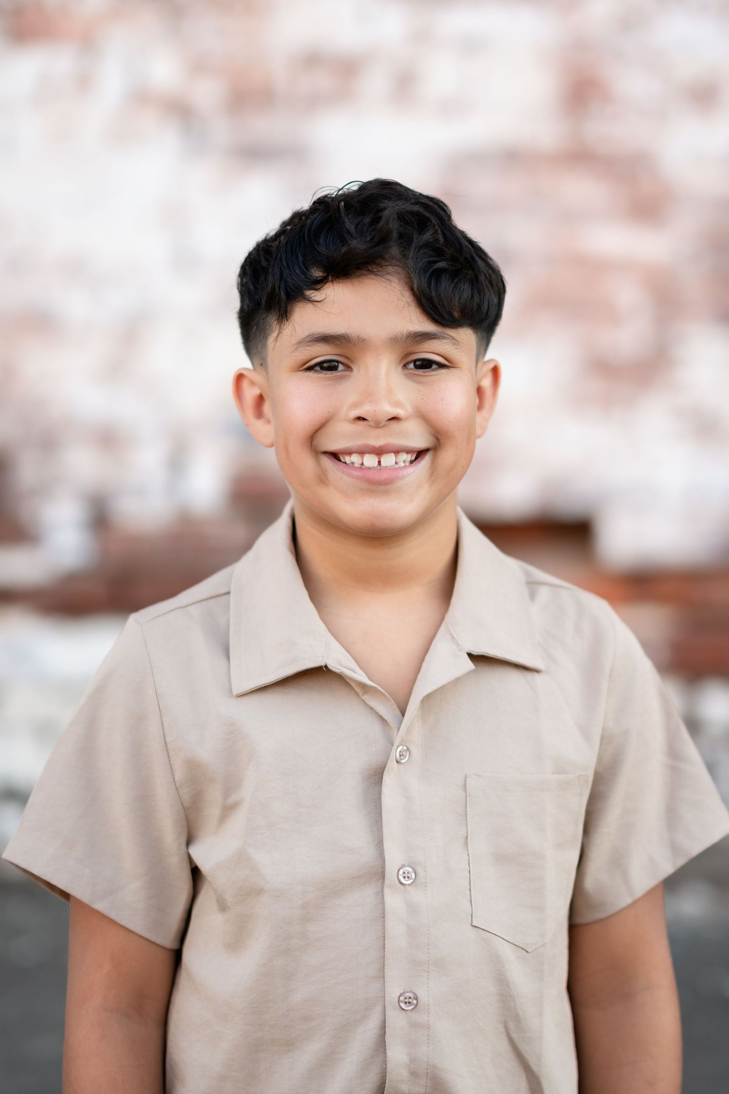 Smiling young boy with dark curly hair wearing a beige short-sleeve button-up shirt, standing against a brick wall.