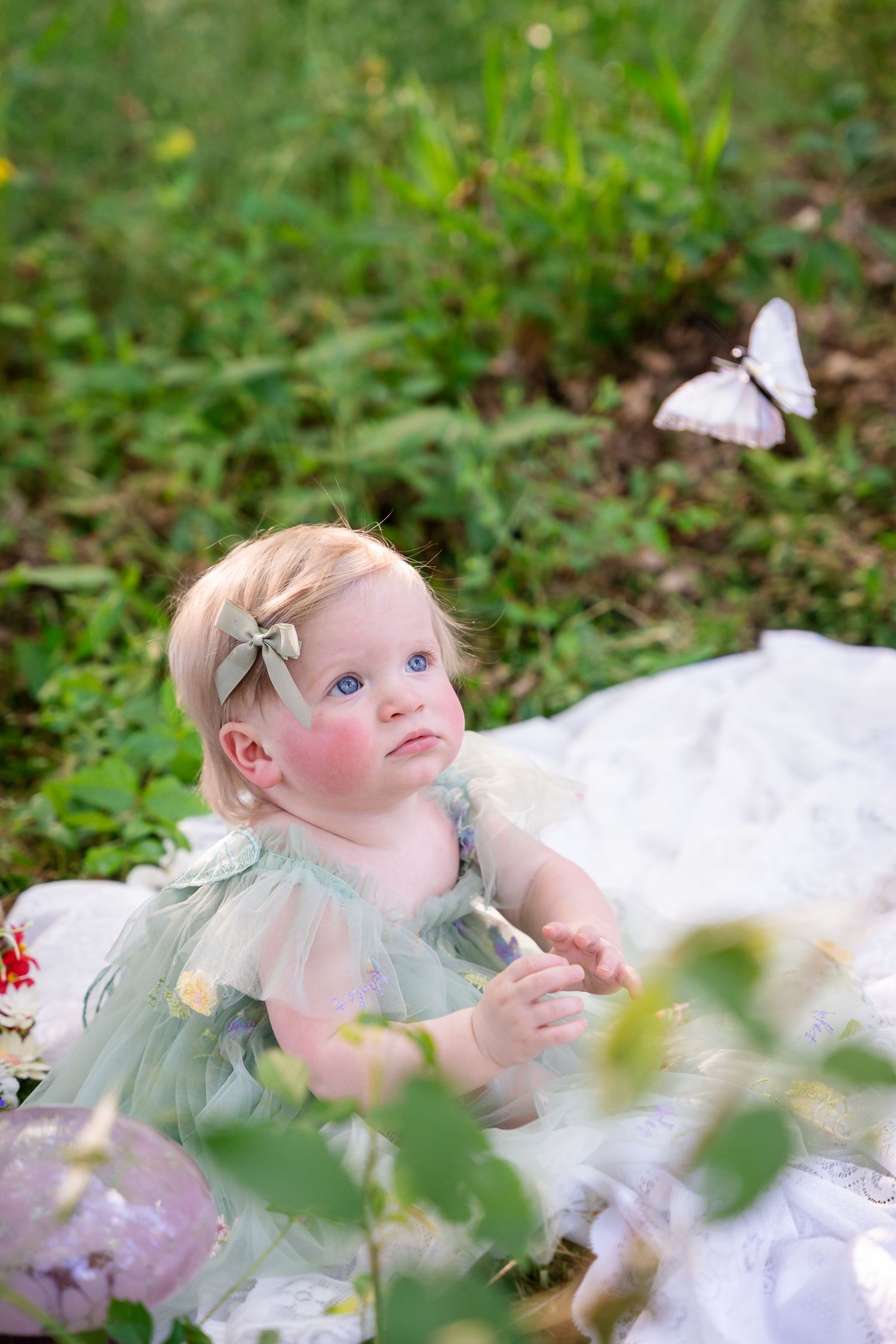 A baby girl in a green tulle dress sits on a white blanket outdoors, gazing at a butterfly in a lush garden.